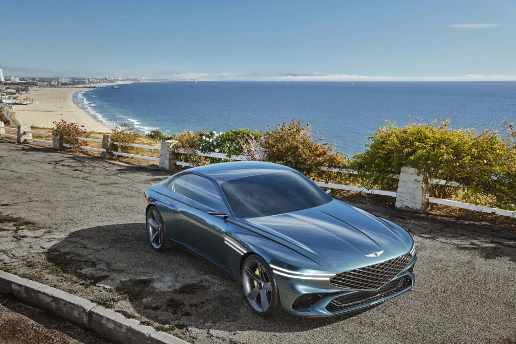 Genesis X Concept 桌布 壁紙 — 一輛時尚金屬藍色跑車停泊在海岸懸崖上，俯瞰著沙灘和廣闊的海洋，天空清澈湛藍。