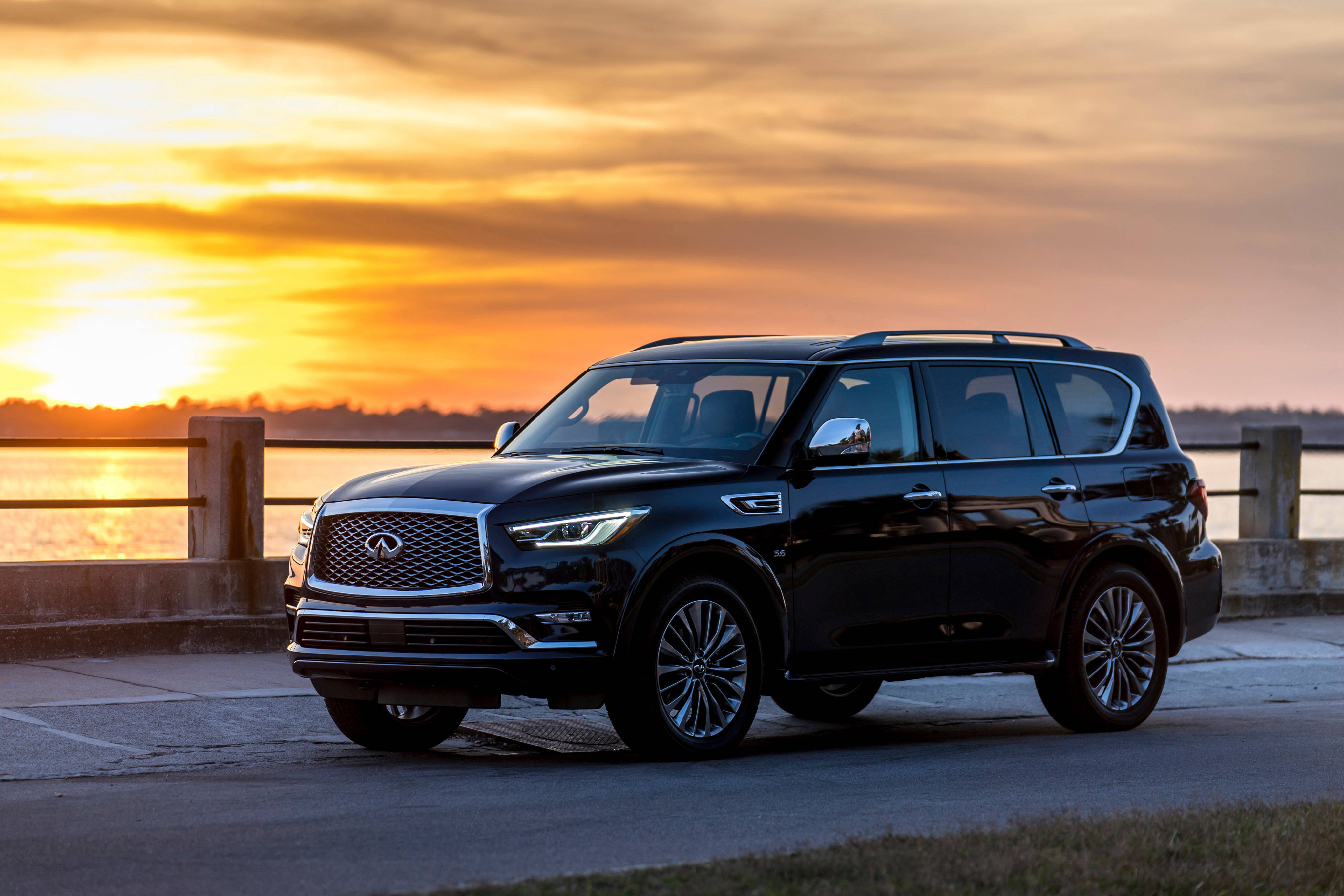 Infiniti 桌布 壁紙 — 一輛時尚黑色 Infiniti QX80 SUV 停泊在海濱碼頭，背景是金色夕陽和溫暖的橙色雲彩。