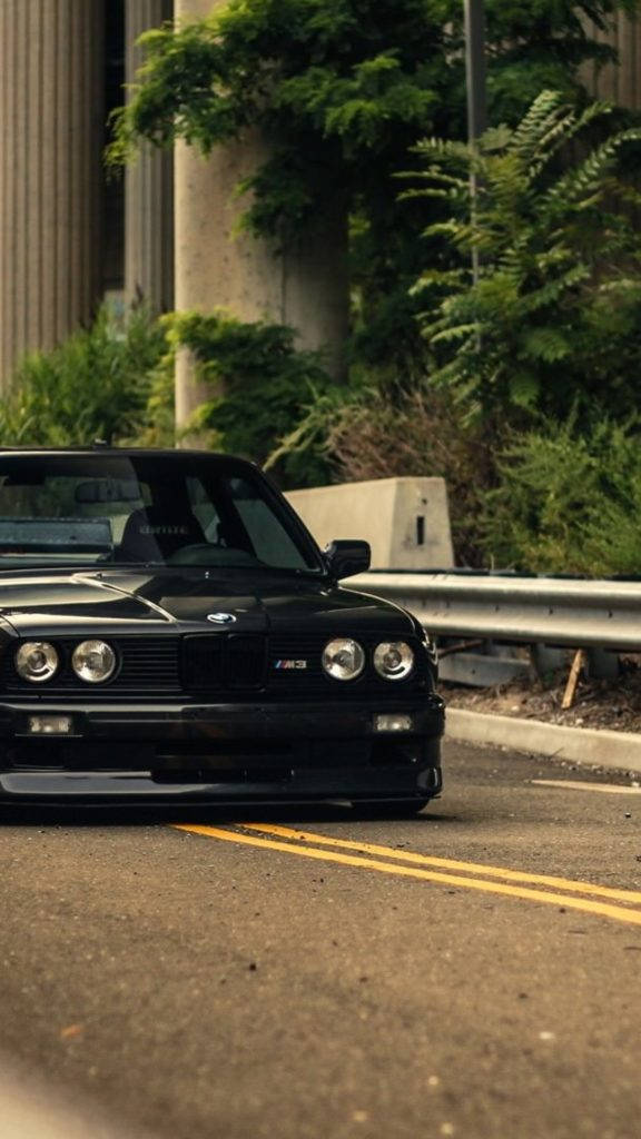 m 桌布 壁紙 — 一輛時尚黑色BMW M3跑車停放在路邊，背景有茂密綠植和混凝土柱。