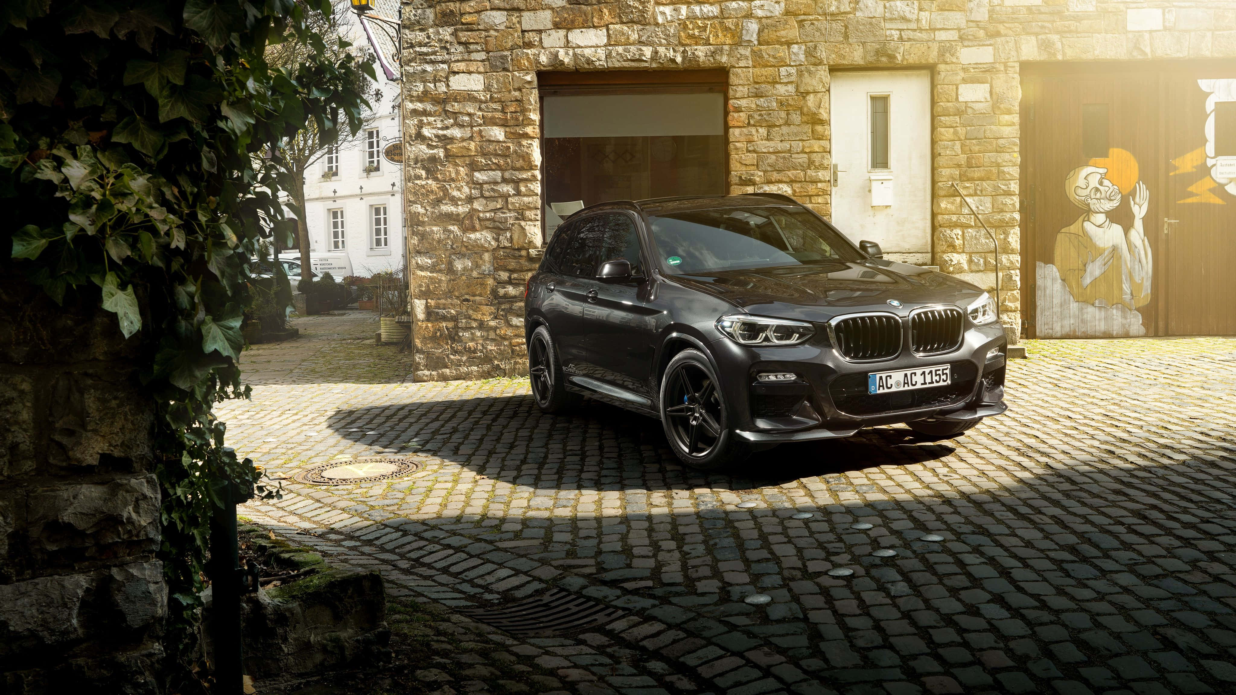 Bmw X3 桌布 壁紙 — 一輛時尚黑色BMW X3 SUV停泊在陽光普照的鵝卵石庭院，旁邊是帶有常春藤和塗鴉藝術的石建築。
