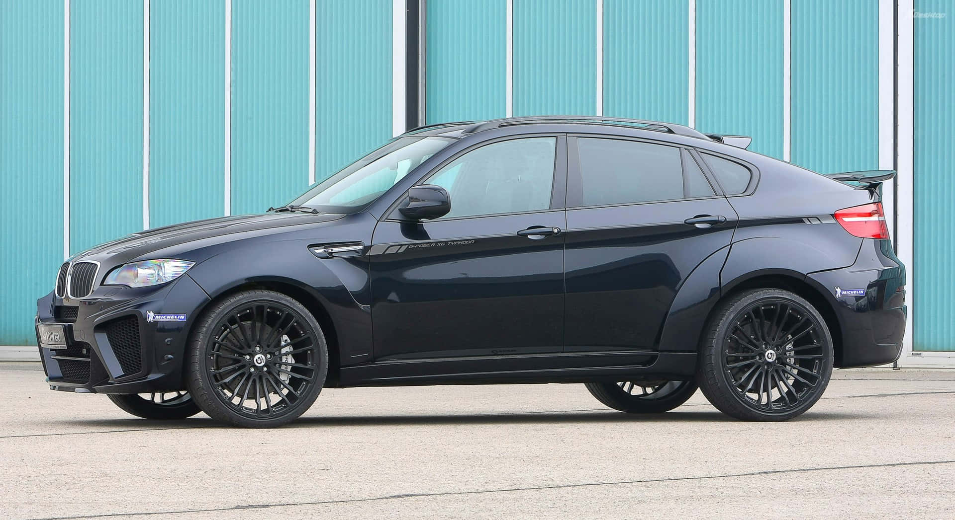 BMW X6 M 桌布 壁紙 — 一輛時尚黑色BMW X6M跑車停放在青綠色波紋金屬牆前。