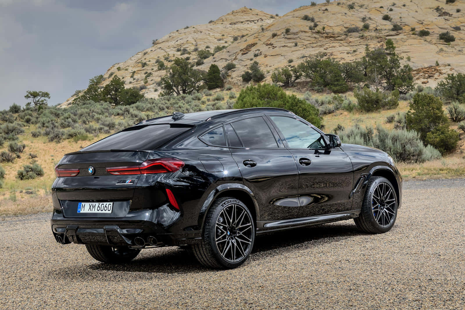 BMW X6 M 桌布 壁紙 — 一輛時尚黑色BMW XM SUV停在碎石上，背景是乾旱的山丘和稀疏的綠植，天空多雲。