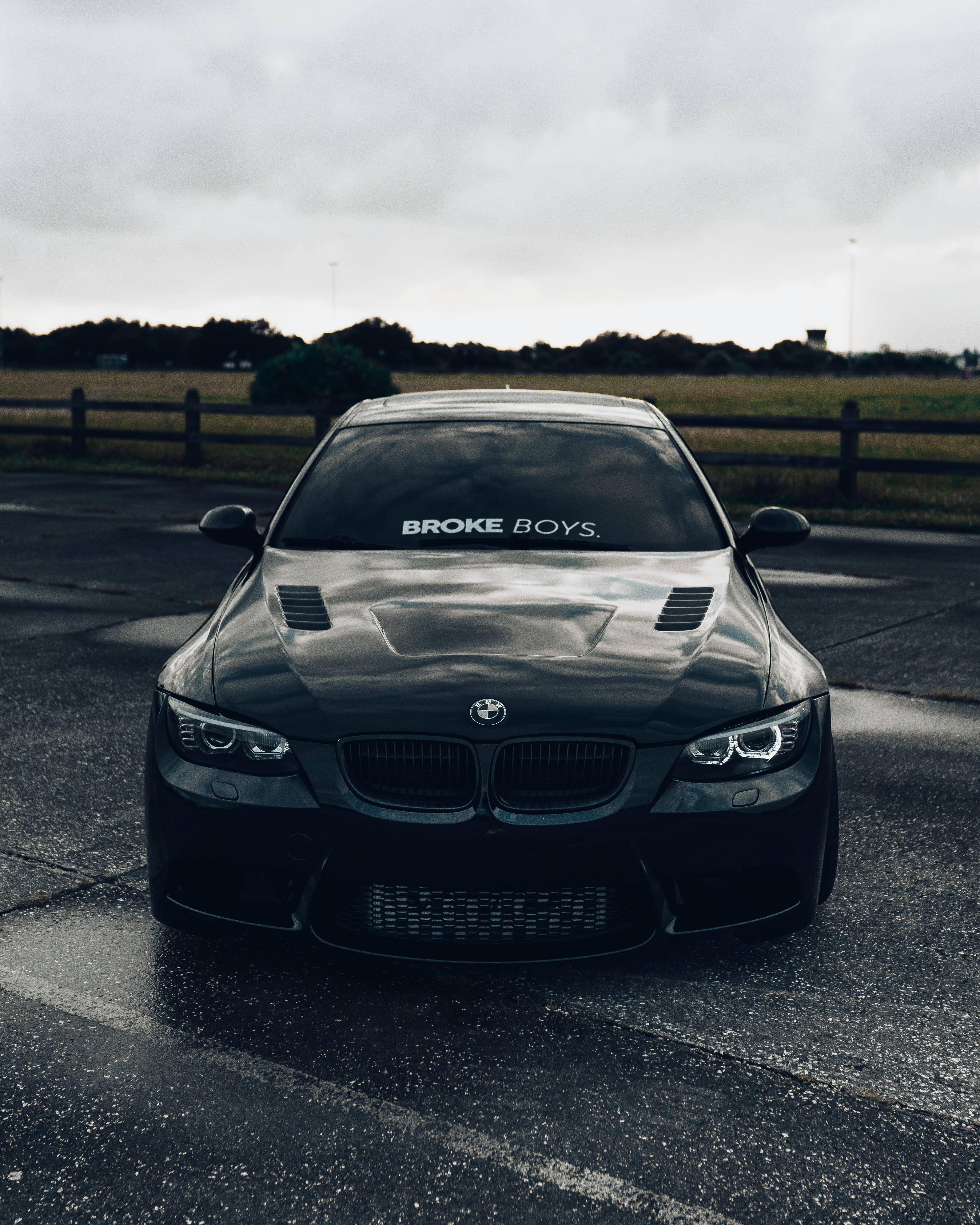 Black Bmw 桌布 壁紙 — 一輛時尚黑色BMW跑車，擋風玻璃上貼有「BROKE BOYS」貼紙，停放在潮濕的瀝青路上，天空多雲。