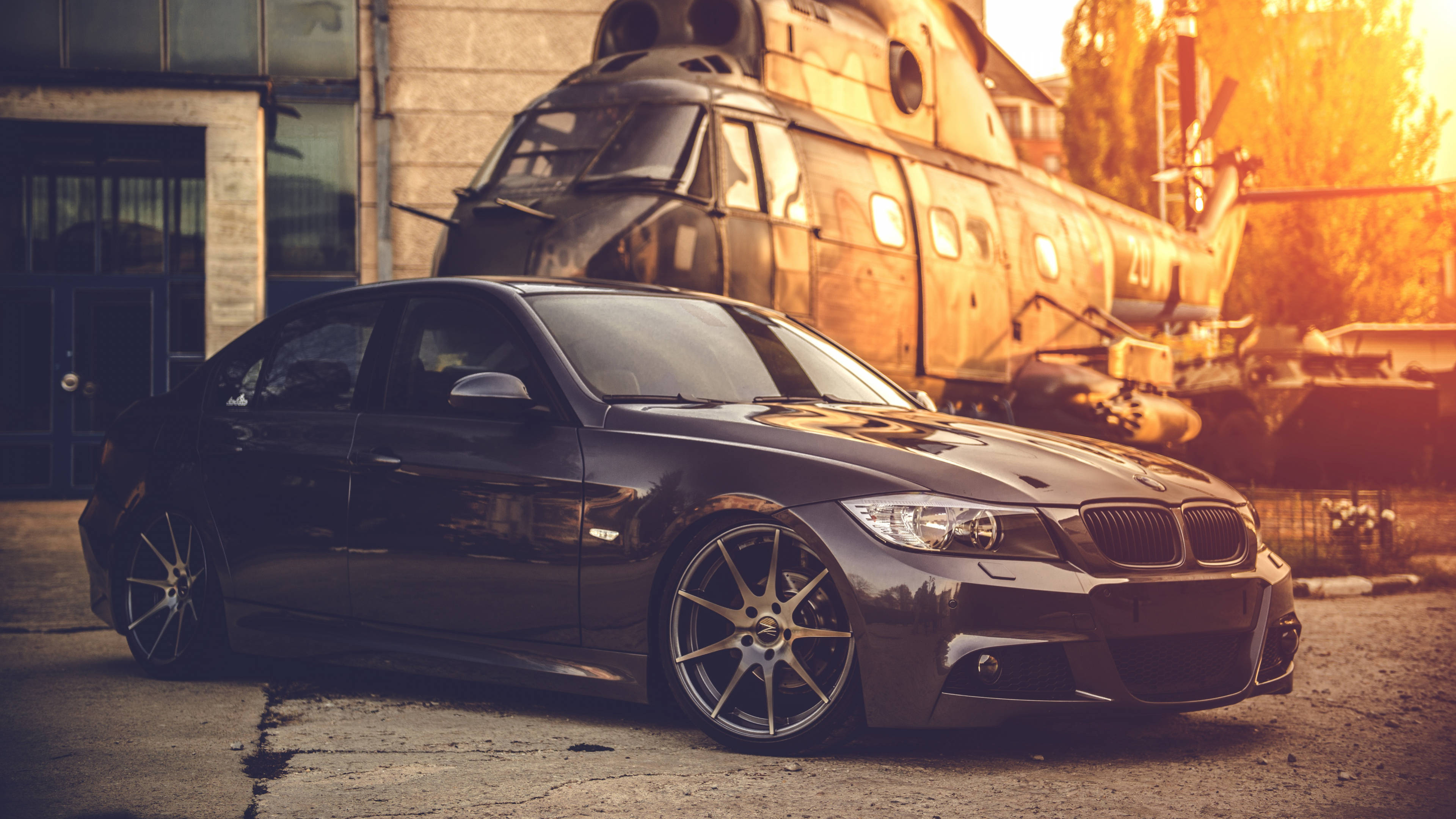4K BMW 壁紙 桌布 壁紙 — 一輛時尚黑色BMW轎車配備訂製輪圈，在溫暖的金色光線下停在一架生鏽的直升機前。