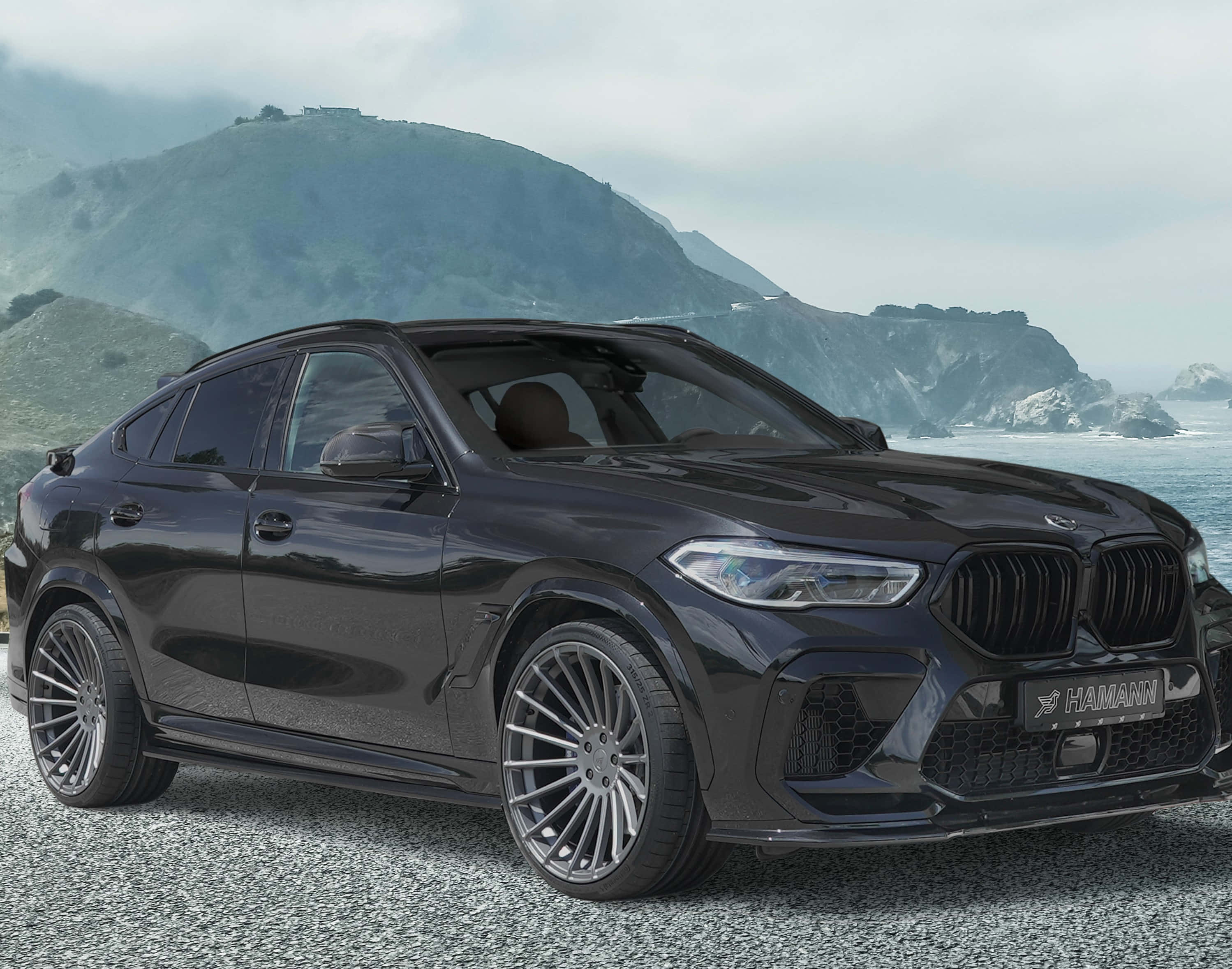 BMW X6 M 桌布 壁紙 — 一輛時尚黑色Hamann改裝BMW X6停在海邊碎石上，背景是霧濛濛的綠色山丘。