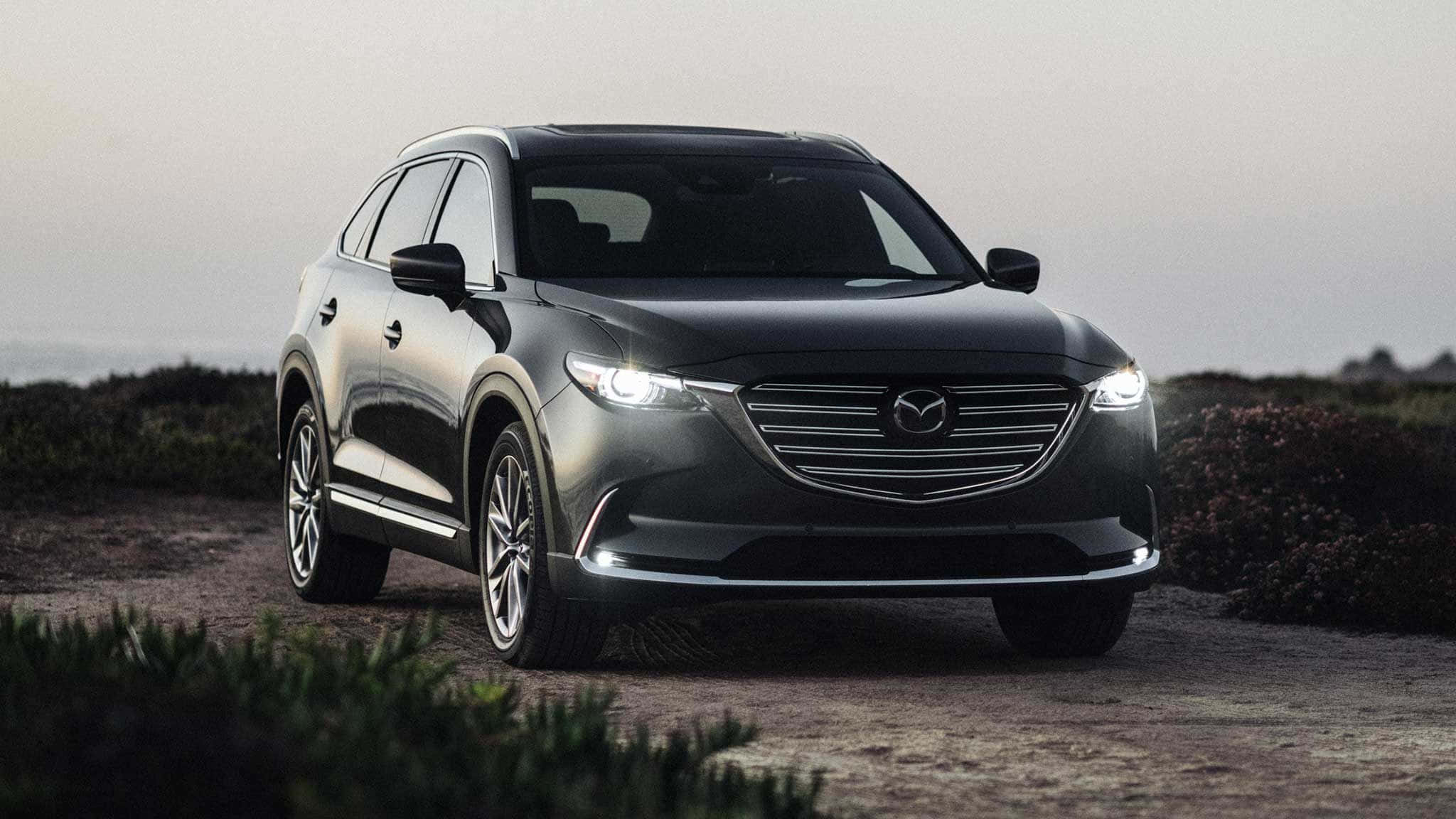 Mazda Cx-9 桌布 壁紙 — 一輛時尚黑色Mazda CX-9 SUV，亮著車頭燈，停泊在塵土小徑旁的低矮灌木叢邊，天空朦朧。