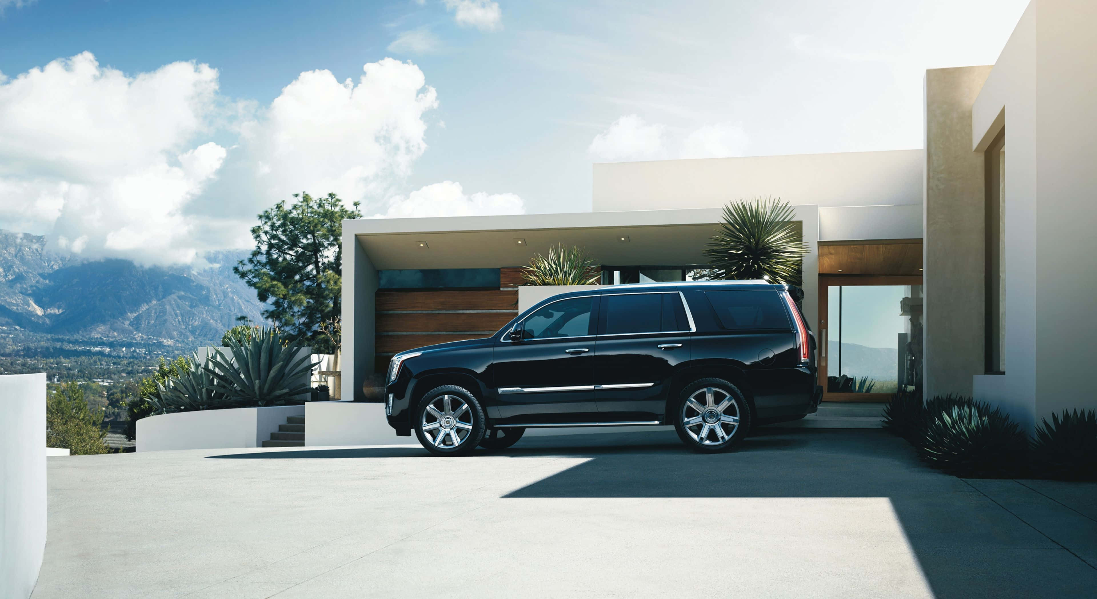 Cadillac Escalade 桌布 壁紙 — 一輛時尚黑色SUV停在現代白色房屋前，背景是山脈和藍天。