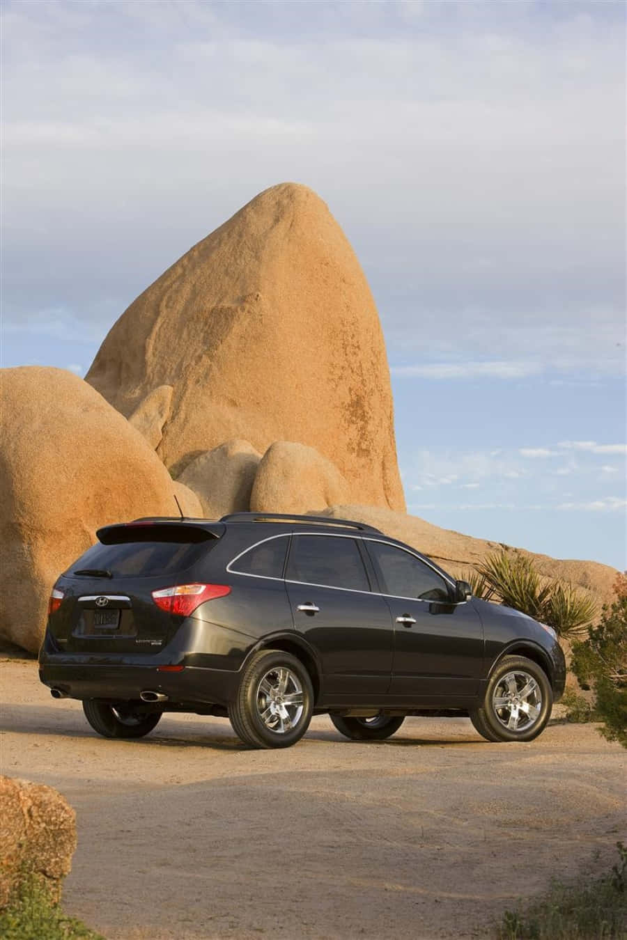 Hyundai Veracruz 桌布 壁紙 — 一輛時尚黑色SUV停泊在塵土飛揚的小徑上，旁邊是巨大的棕褐色巨石，柔和藍天中散佈著雲朵。