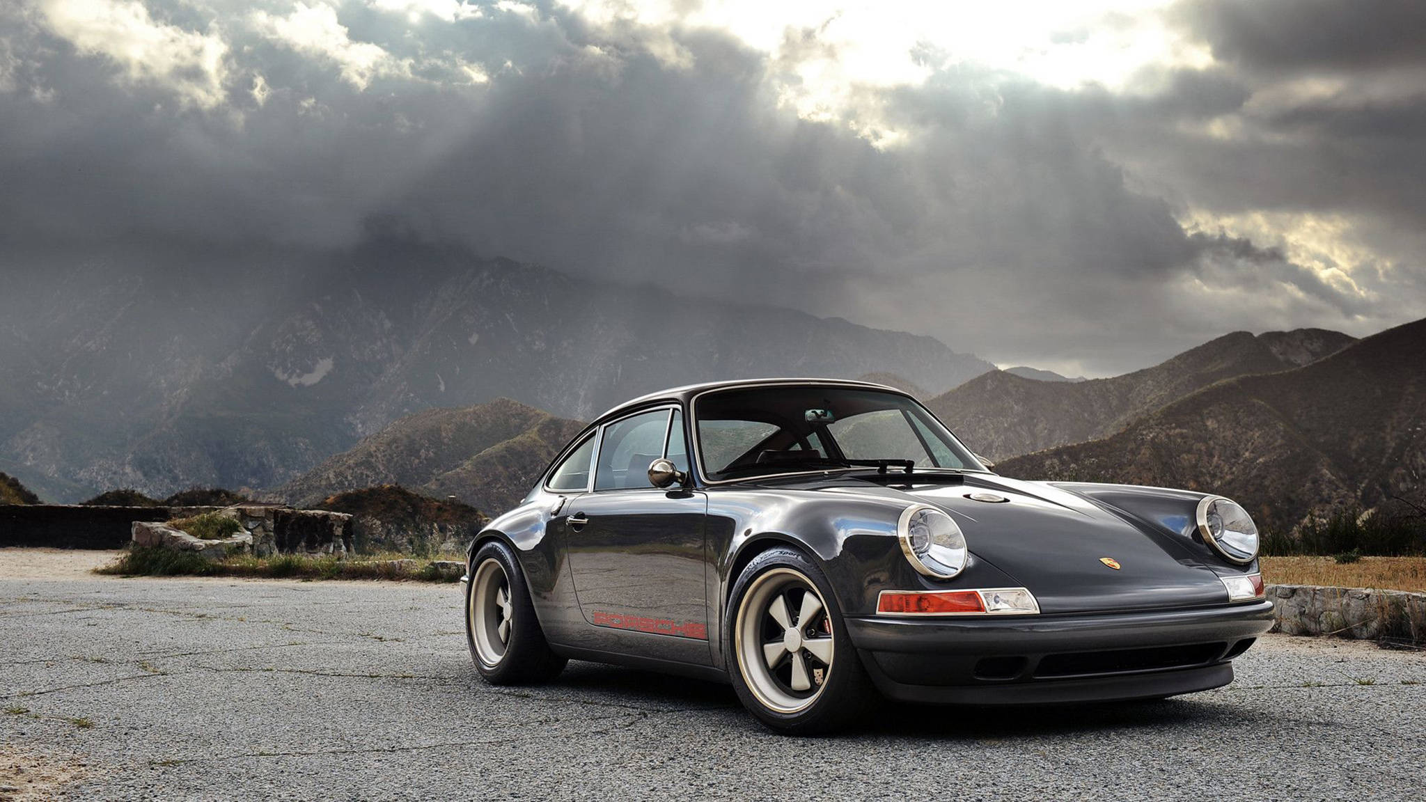 Singer 桌布 壁紙 — 一輛時尚黑色保時捷911跑車停泊在碎石路上，背景是風暴天空下的戲劇性山脈。