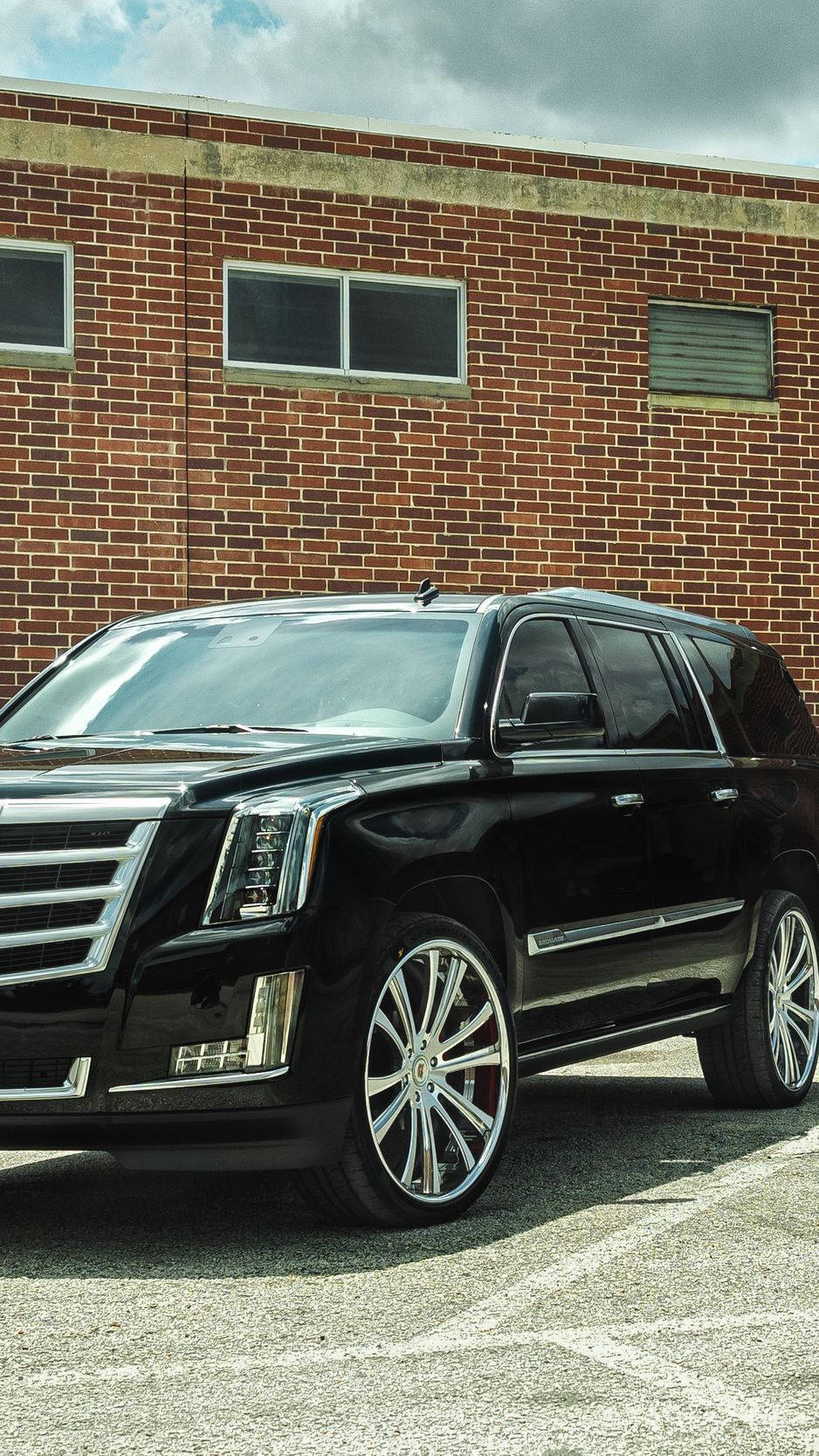 凱迪拉克 iPhone 桌布 壁紙 — 一輛時尚黑色凱迪拉克 Escalade，配備訂製鍍鉻輪圈，停放在紅磚建築前，天空多雲。