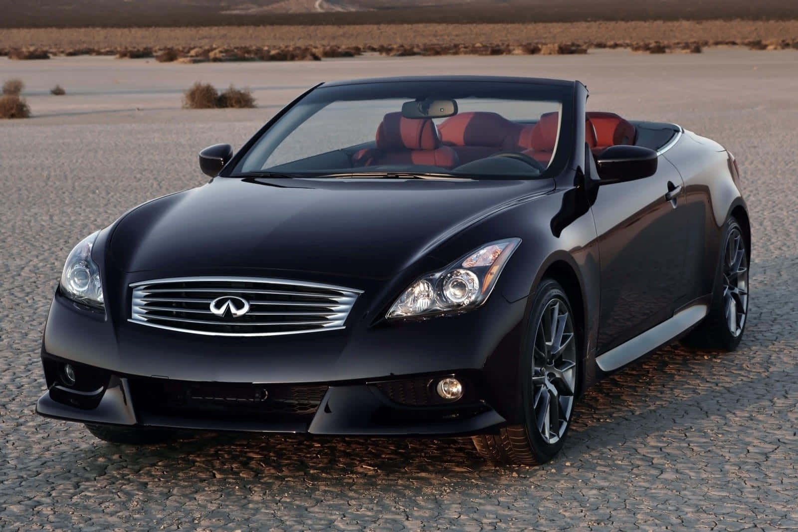Infiniti Q60 桌布 壁紙 — 一輛時尚黑色敞篷跑車搭配紅色內裝，停放在溫暖金色光線下龜裂的沙漠道路上。