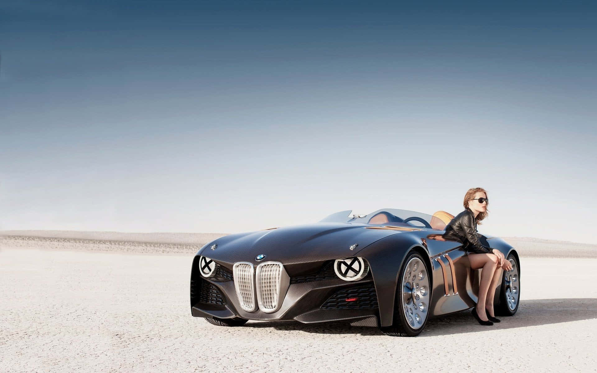 Bmw 328 桌布 壁紙 — 一輛時尚黑色概念車，一位戴太陽眼鏡的女士在晴朗天空下的廣闊沙漠平原上靠在車旁邊休息。