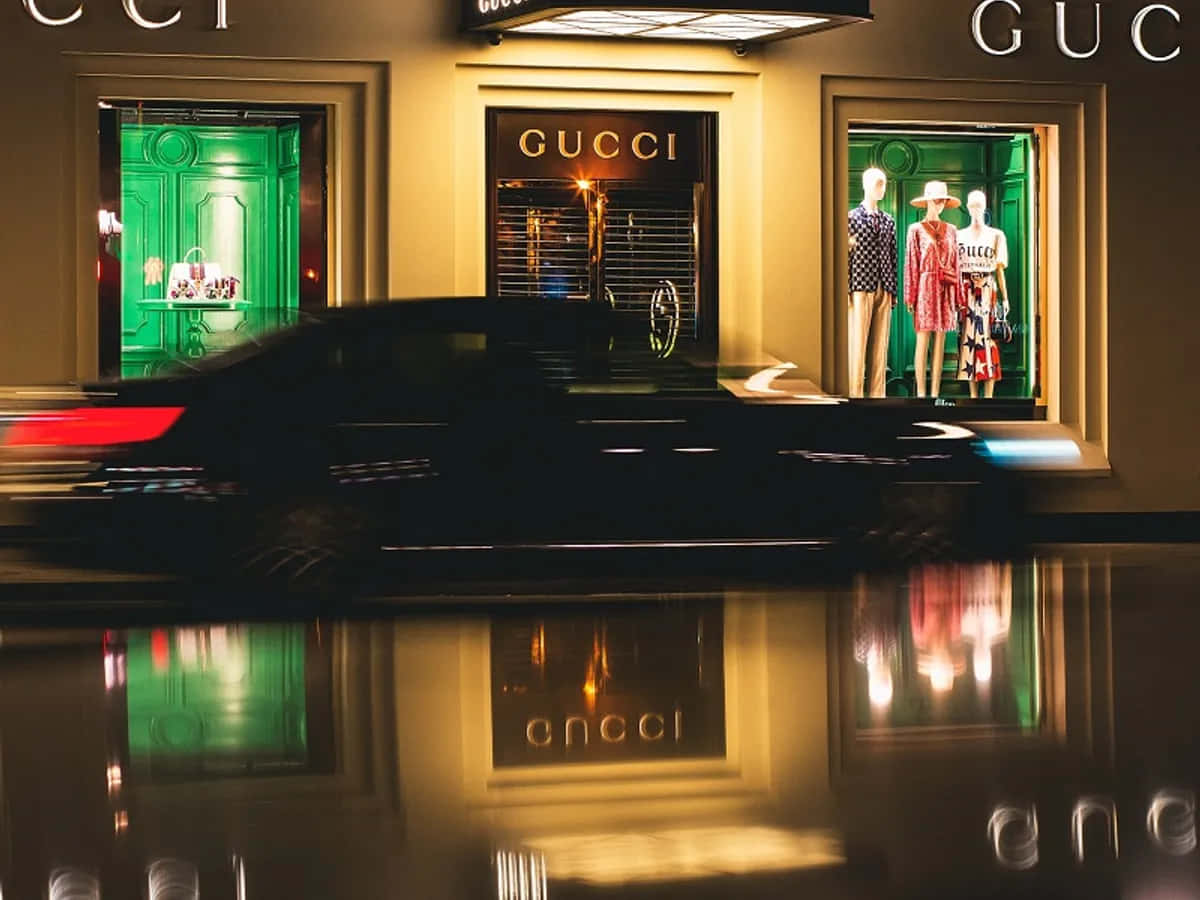 Gucci 桌布 壁紙 — 一輛時尚黑色汽車在夜間駛過Gucci商店，其霓虹綠色窗戶和金色招牌映照在濕漉漉的人行道上。