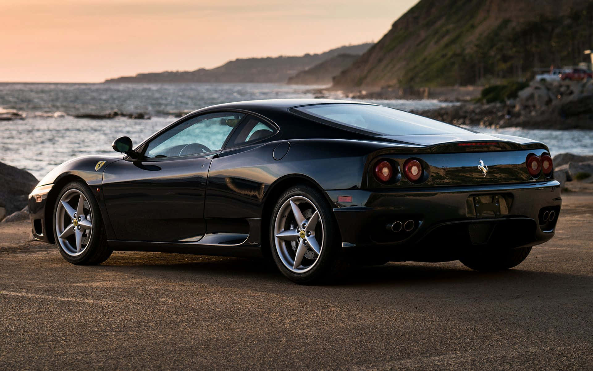 Ferrari 360 Modena 桌布 壁紙 — 一輛時尚黑色法拉利停泊在日落時分的海岸公路上，背景是海洋和懸崖。