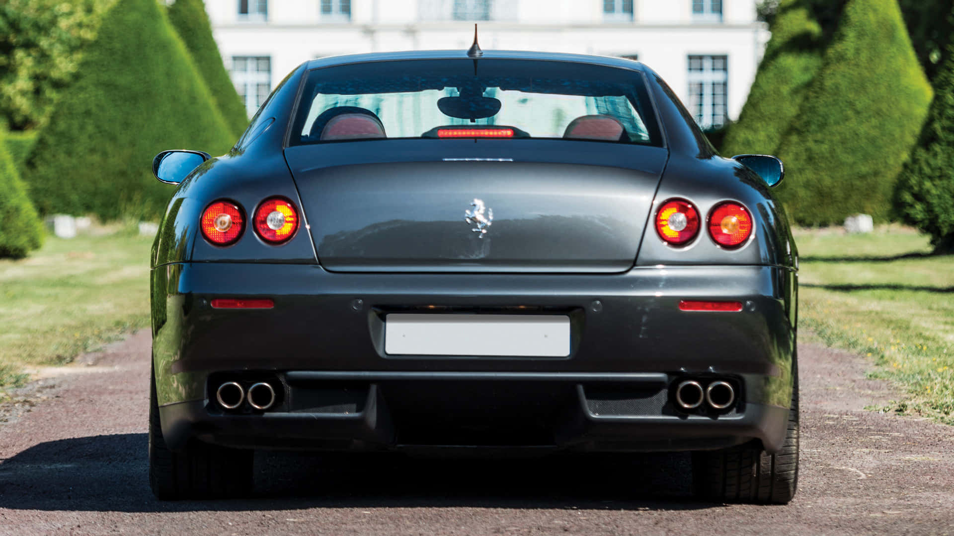 Ferrari 612 Scaglietti 桌布 壁紙 — 一輛時尚黑色法拉利停泊在碎石路上，兩旁是修剪整齊的樹籬和一座宏偉的白色大宅的後視圖。
