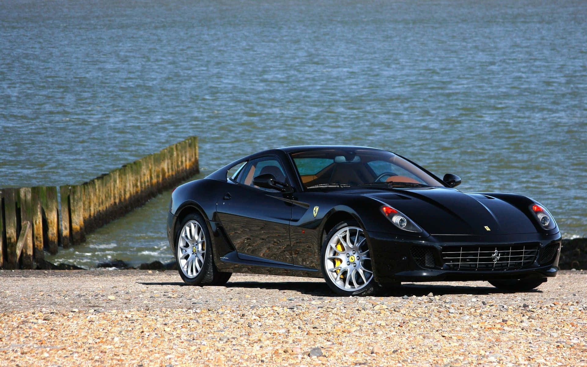 Ferrari 599 Gtb Fiorano 桌布 壁紙 — 一輛時尚黑色法拉利跑車停泊在平靜水域旁邊的卵石灘上，附近有木樁。