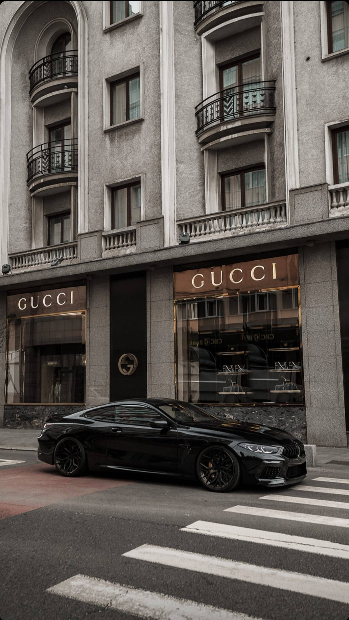 Gucci iPhone 桌布 壁紙 — 一輛時尚黑色跑車停在城市街道上，前面是裝飾華麗的Gucci商店。