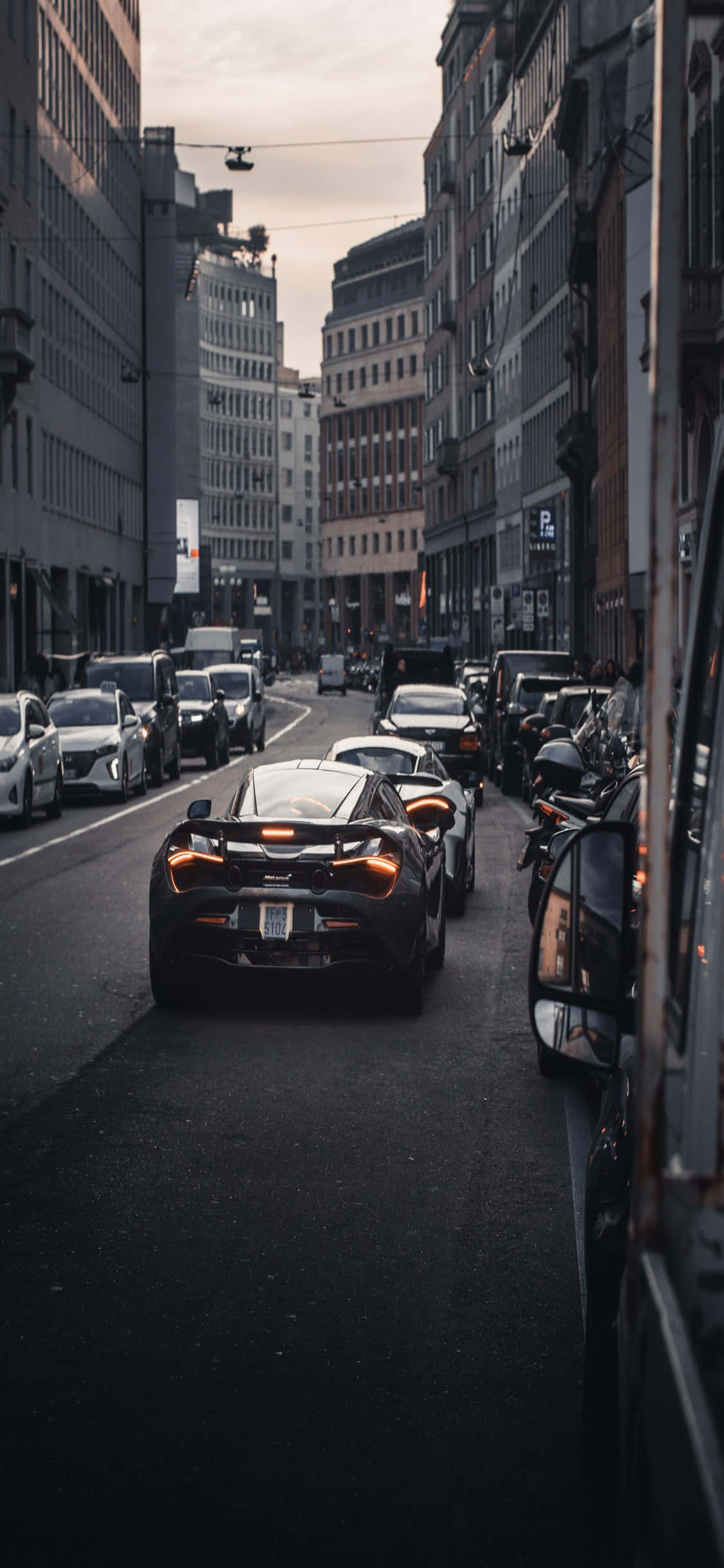 Black Car 桌布 壁紙 — 一輛時尚黑色跑車在暮色蒼茫的天空下，行駛在兩旁高樓林立的狹窄城市街道上。