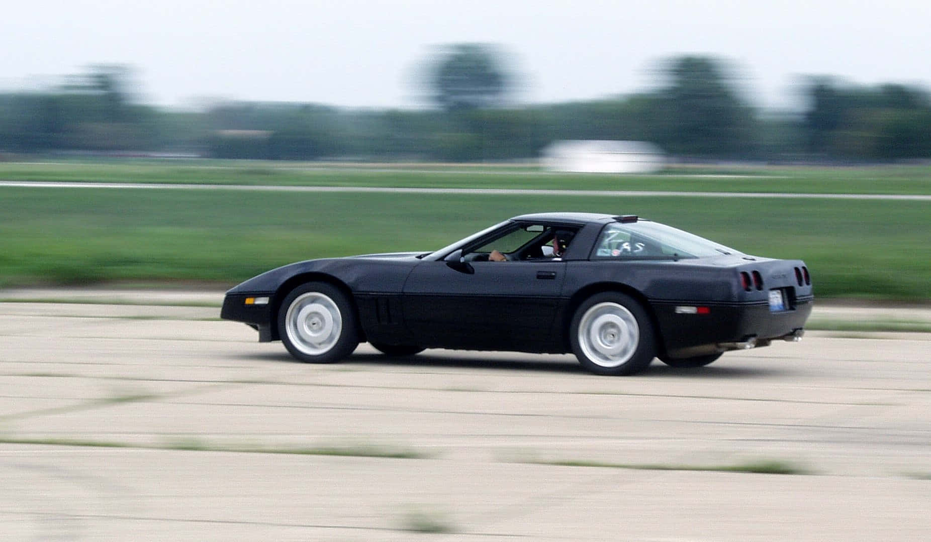 C4 Corvette 桌布 壁紙 — 一輛時尚黑色跑車在鋪砌的賽道上疾馳，背景是模糊的綠植。