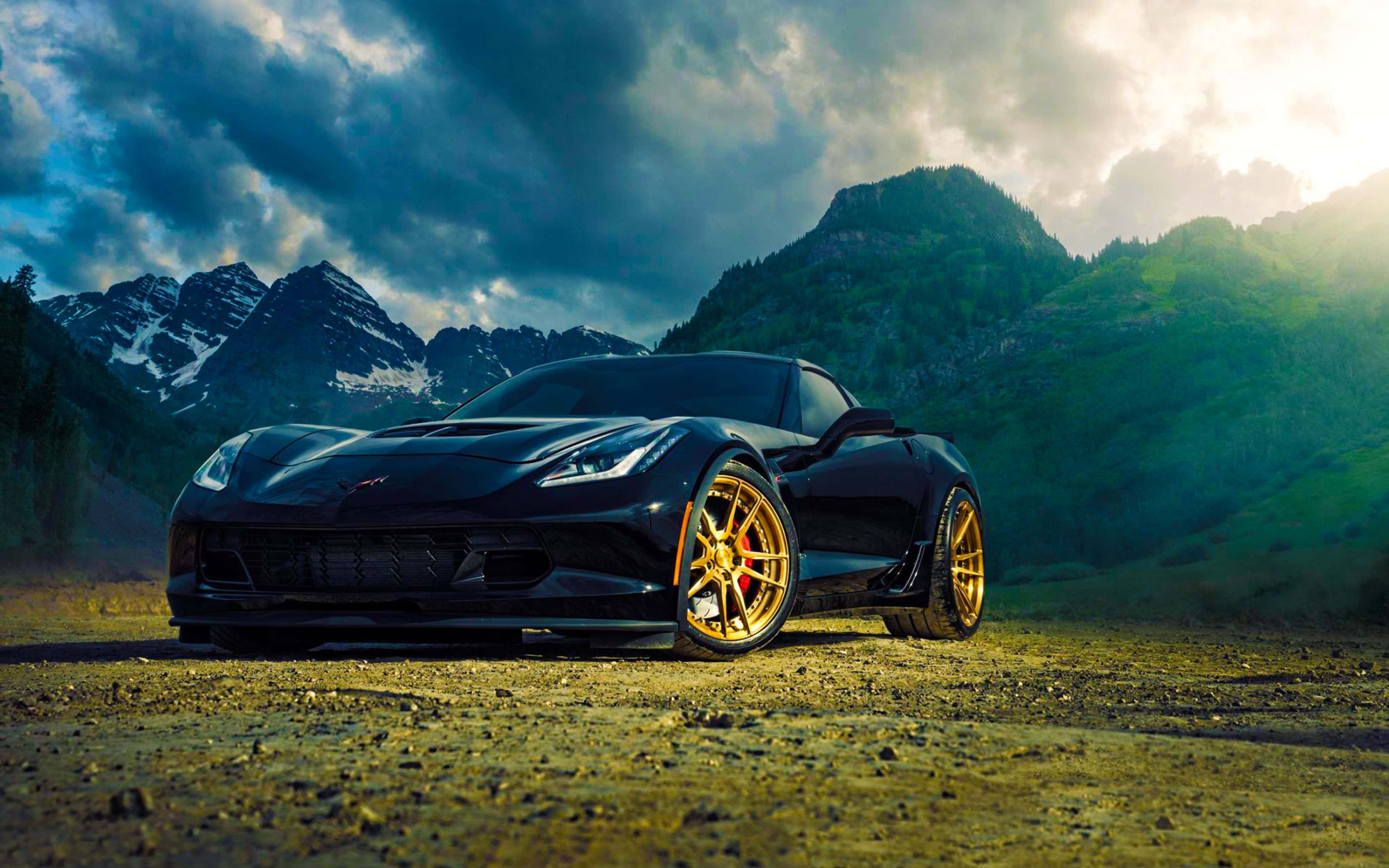 Chevrolet Corvette Z06 桌布 壁紙 — 一輛時尚黑色跑車搭配金色輪圈，停泊在碎石路上，背景是白雪覆蓋的山脈和戲劇性的雲層。