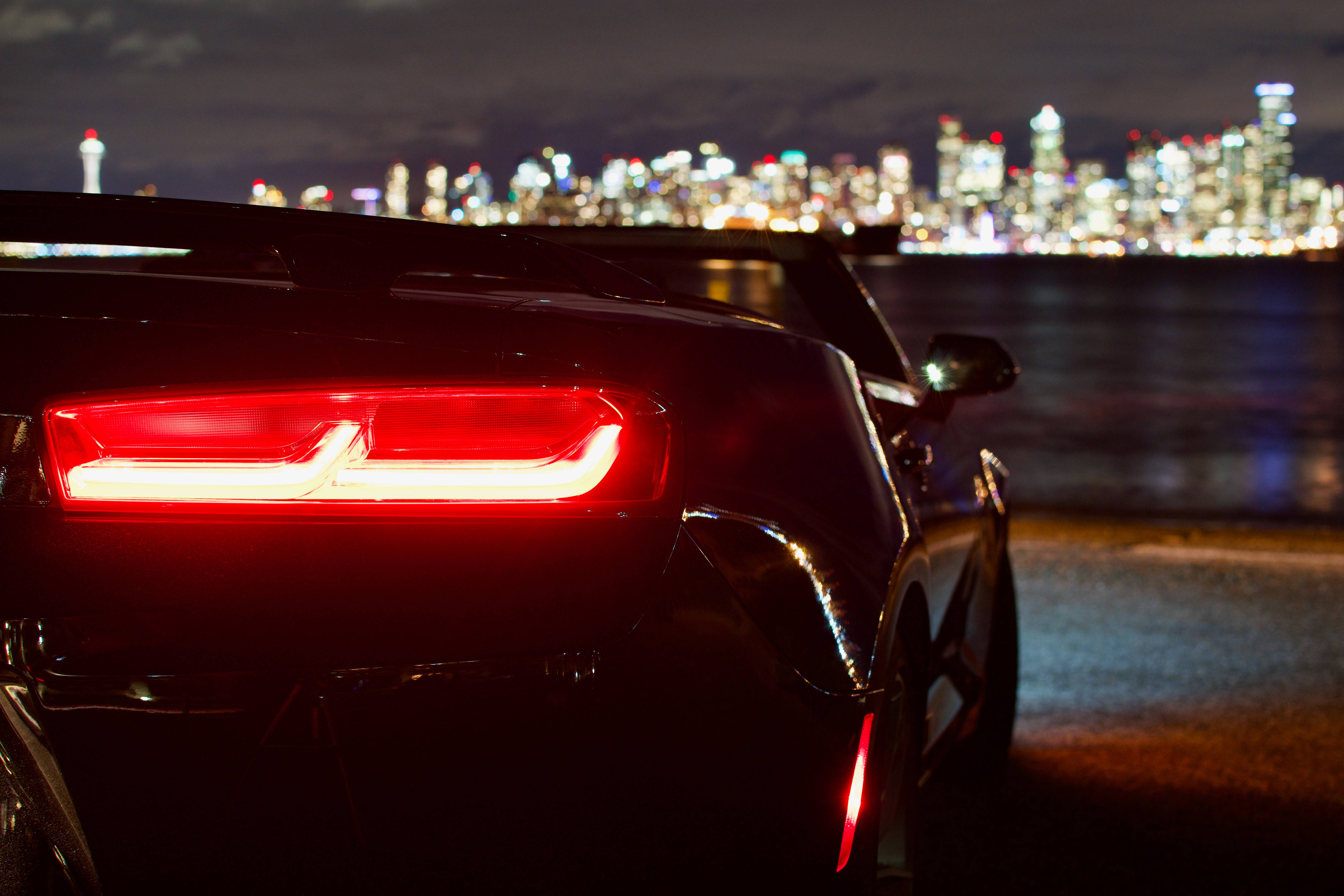 Chevrolet 桌布 壁紙 — 一輛時尚黑色跑車的發光紅色尾燈在夜間城市天際線倒映在水面上的背景下格外突出。