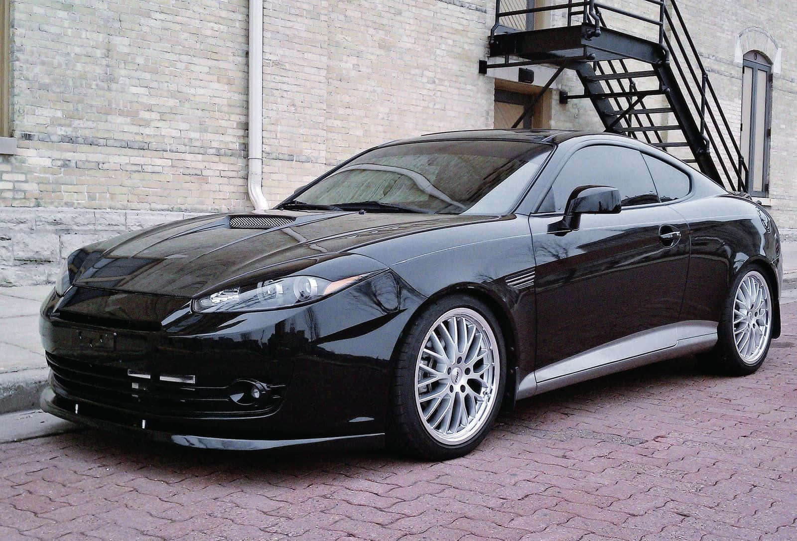 Hyundai Tiburon 桌布 壁紙 — 一輛時尚黑色跑車配銀色輪圈，停在紅磚人行道上，旁邊是米色磚建築物，有黑色金屬樓梯。