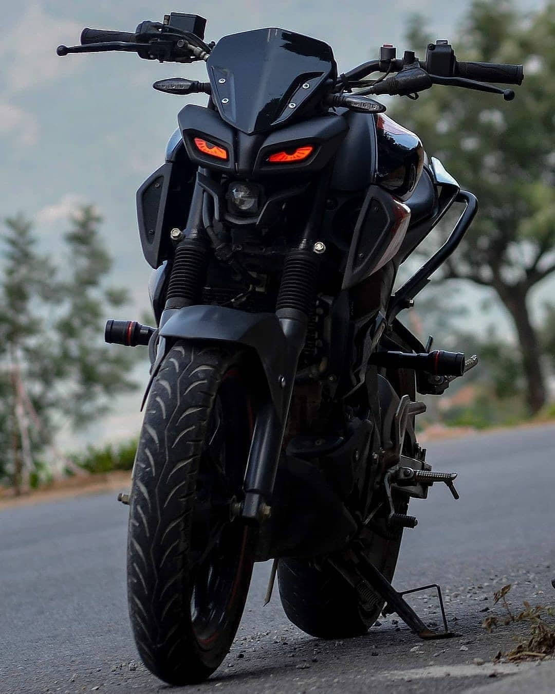 yamaha mt 15 桌布 壁紙 — 一輛時尚黑色運動機車，帶有發光的橙色眼睛，停在樹木環繞的柏油路上，天空多雲。