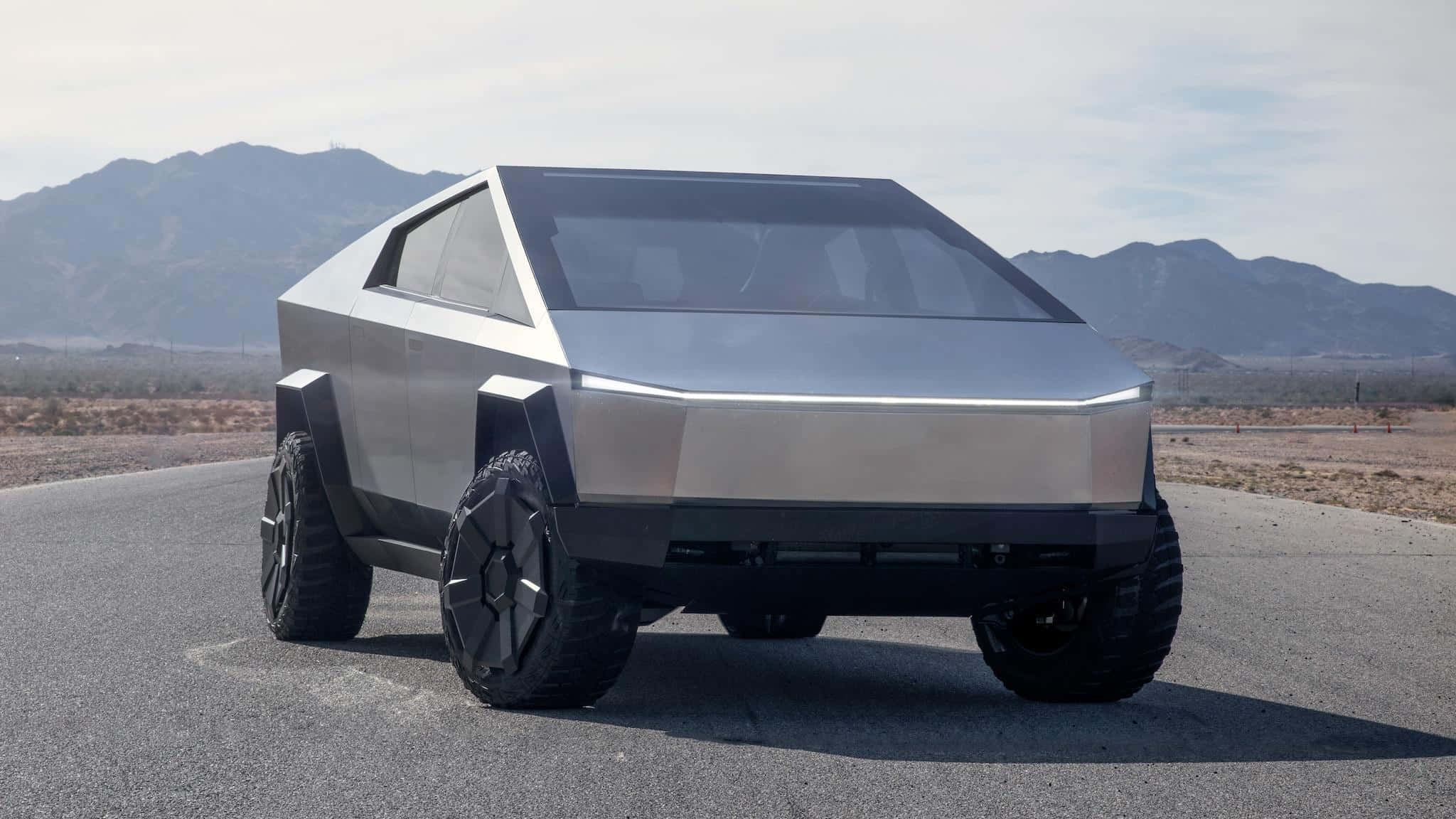 Tesla Cybertruck 桌布 壁紙 — 一輛未來感十足的銀色 Tesla Cybertruck，採用稜角設計，停在沙漠公路上，背景有山脈。