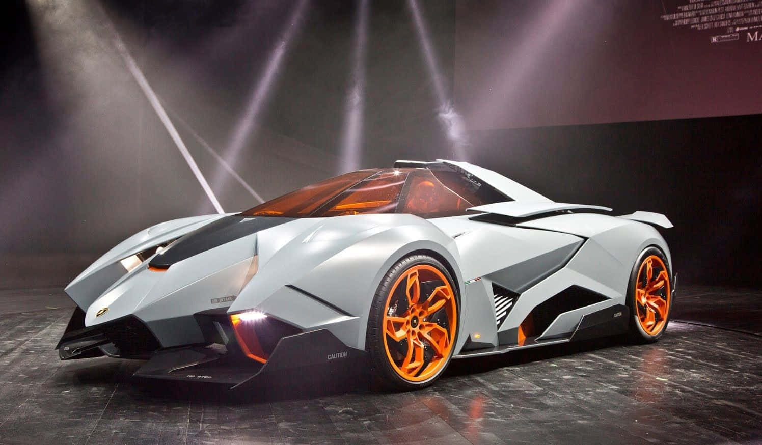 Top 10 Car 桌布 壁紙 — 一輛未來感的銀色藍寶堅尼概念車，配備橙色車輪，在黑暗舞台上被聚光燈戲劇性地照亮。