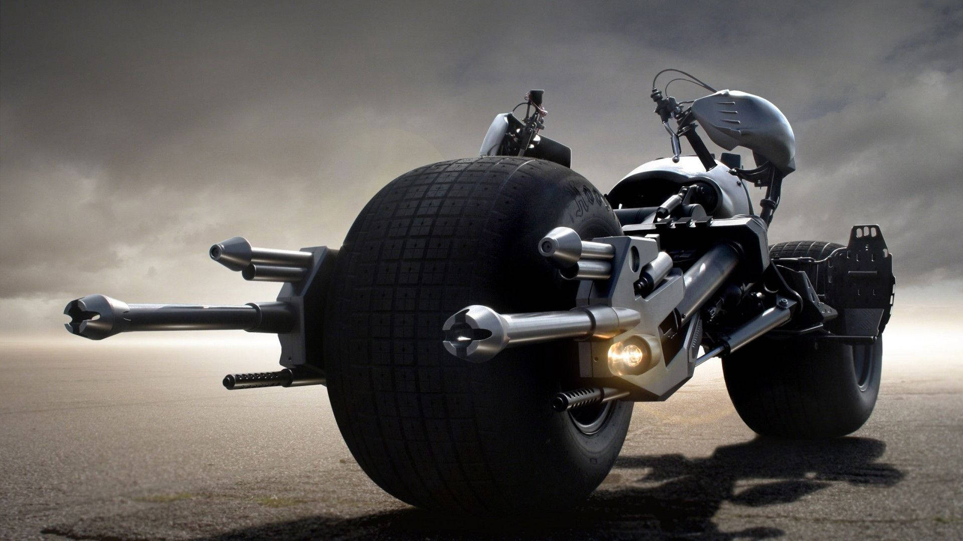 Batmobile 桌布 壁紙 — 一輛未來風格的摩托車，配備巨大輪胎和暴露的機械部件，矗立在暴風雨天空下的荒涼道路上。