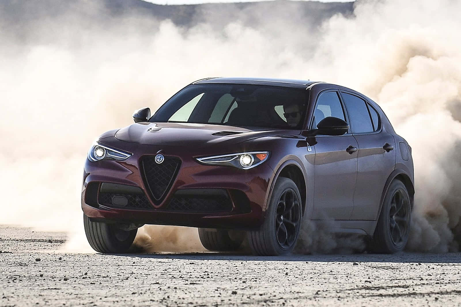 愛快羅密歐 桌布 壁紙 — 一輛栗色Alfa Romeo SUV在塵土飛揚的沙漠景觀中奔馳，揚起沙塵和礫石。