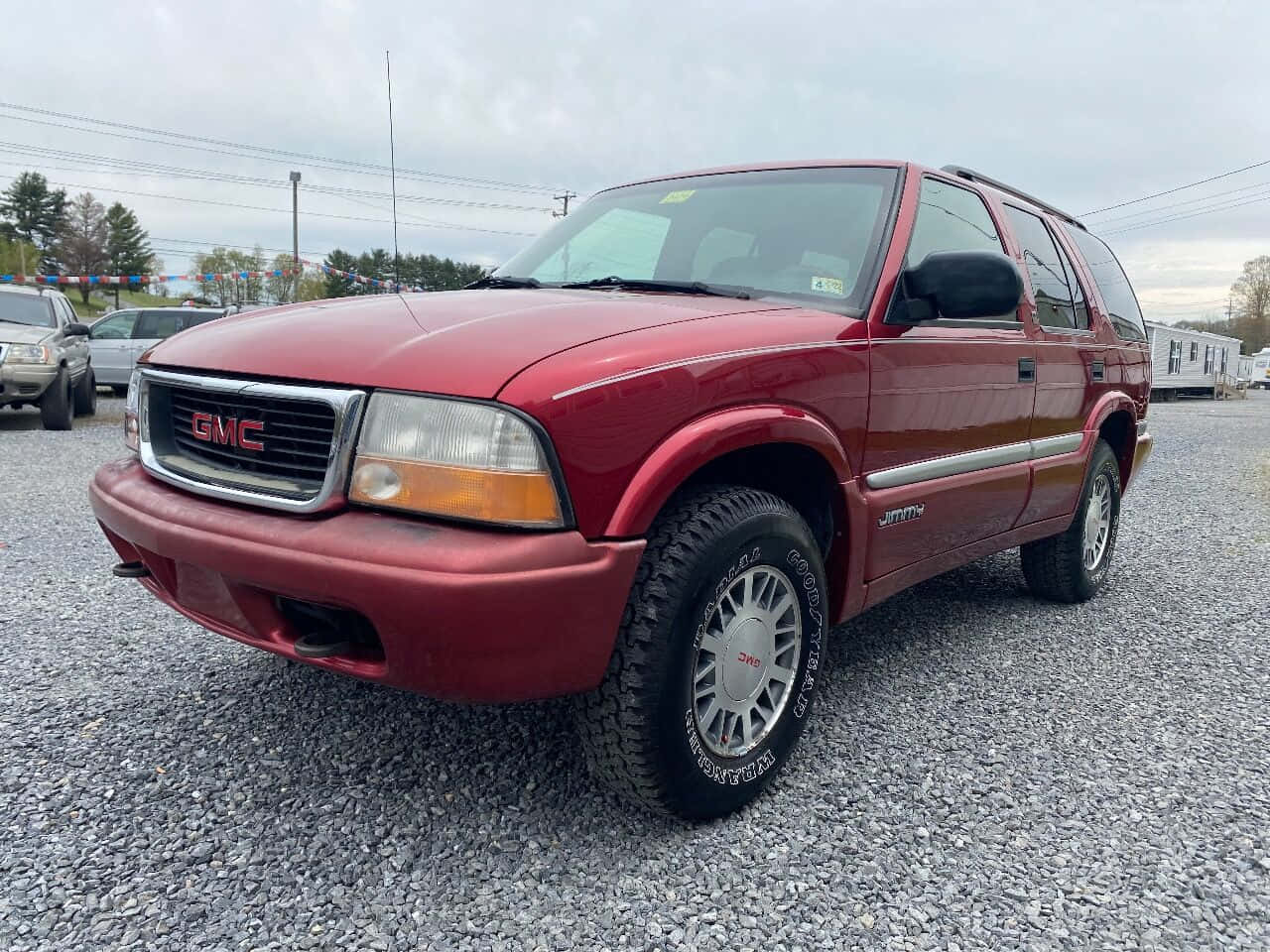 Gmc Jimmy 桌布 壁紙 — 一輛栗色GMC Jimmy SUV停放在碎石上，背景有樹木和陰天。