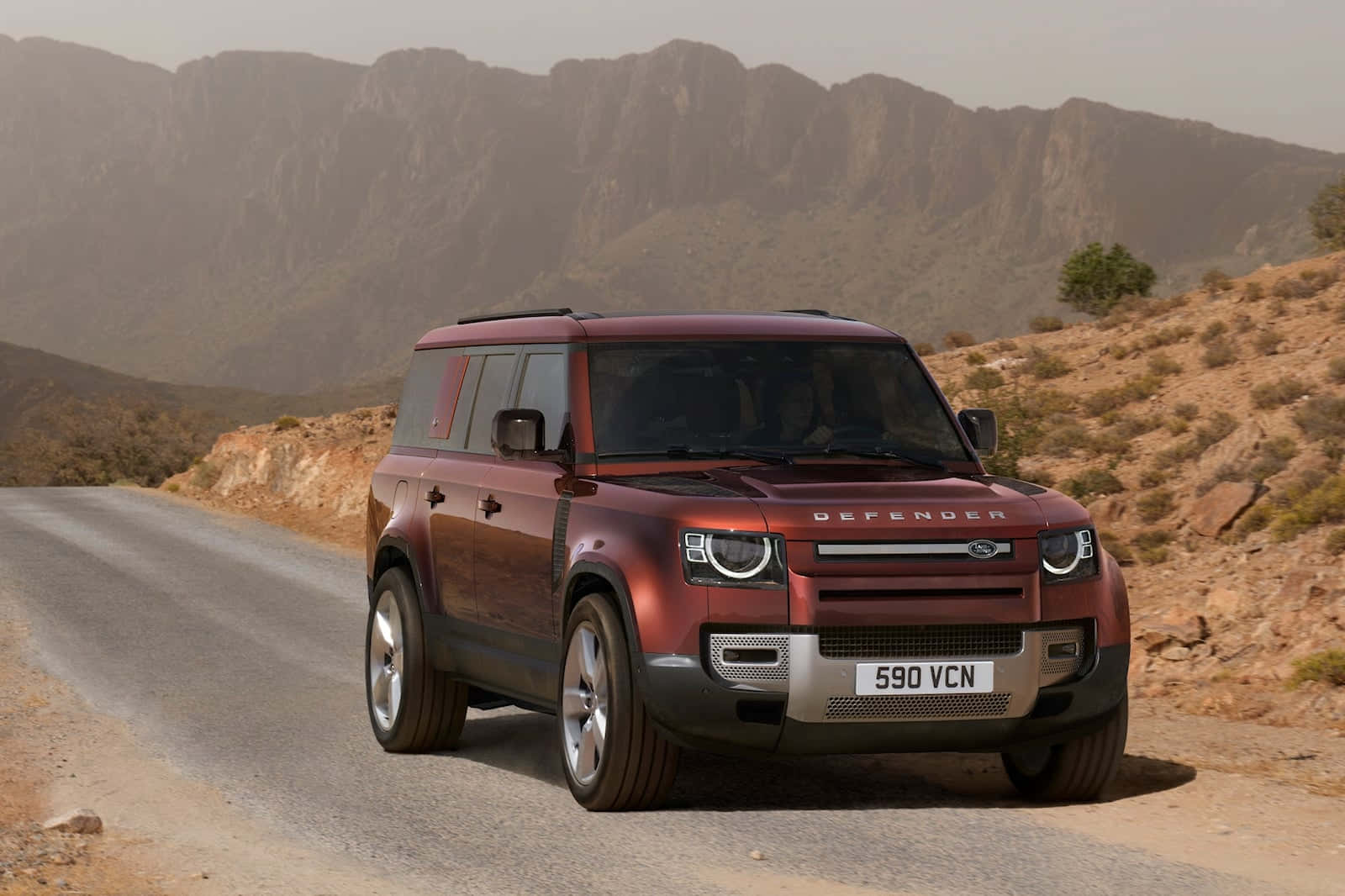 Land Rover Defender 桌布 壁紙 — 一輛栗色Land Rover Defender在朦朧日光下行駛於蜿蜒山路，穿越乾旱地形。