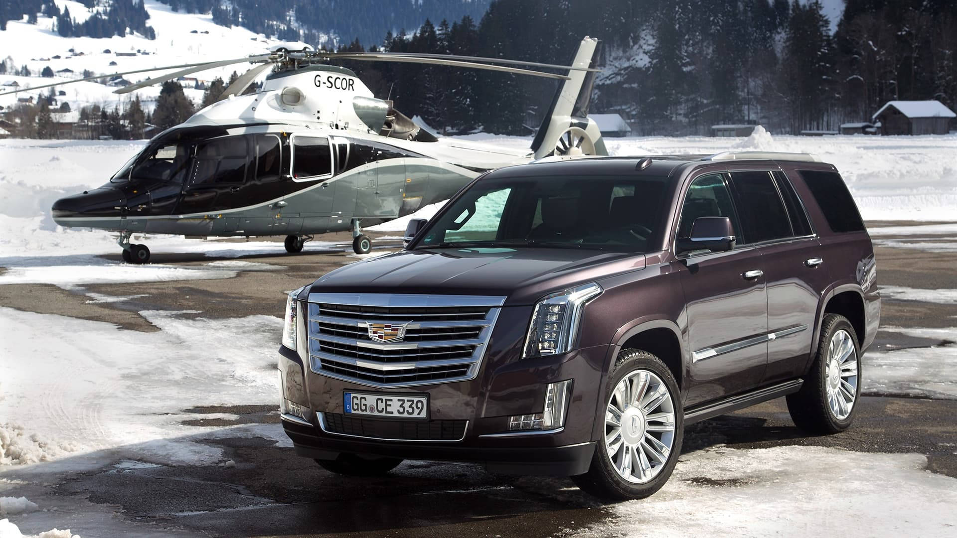 Cadillac 桌布 壁紙 — 一輛栗色凱迪拉克 Escalade SUV 停放在白雪覆蓋的停機坪上，旁邊有一架黑白直升機，背景是冬季山景。