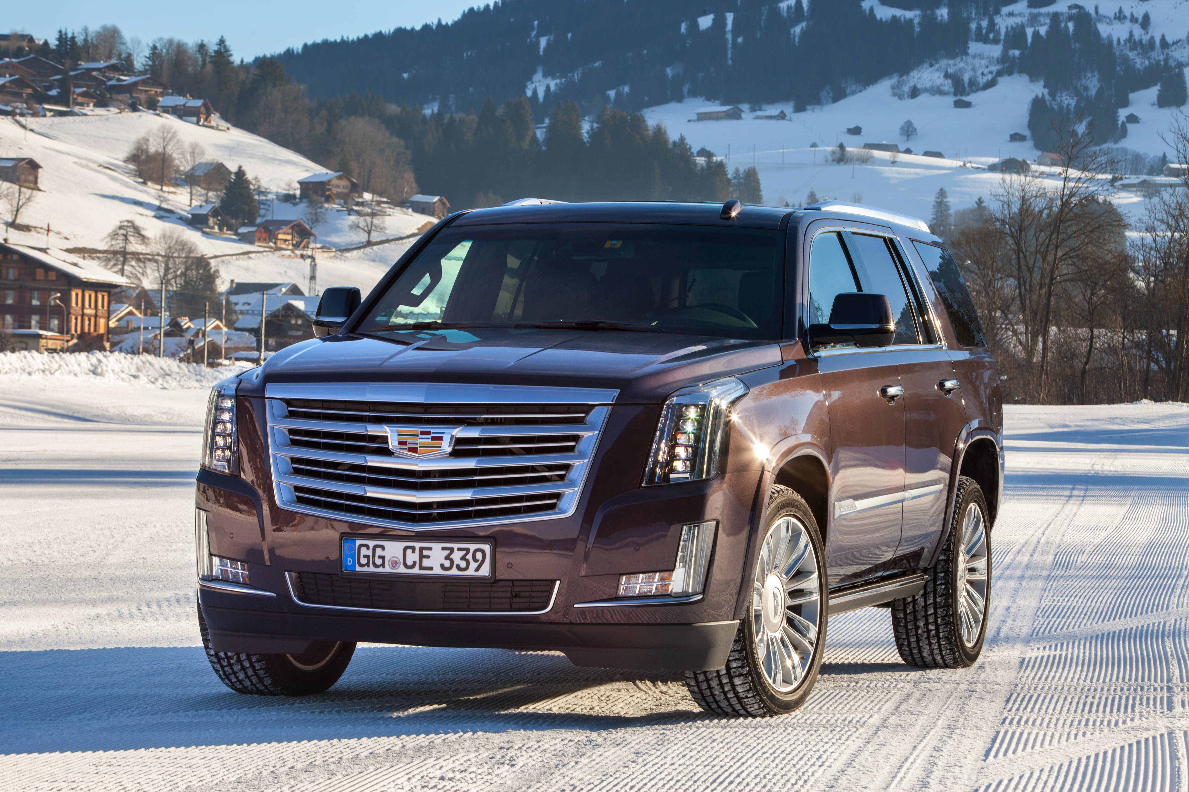 cadillac iPhone 桌布 壁紙 — 一輛栗色凱迪拉克SUV在雪路上行駛，穿過風景如畫的阿爾卑斯山村，背景是白雪覆蓋的山脈和木屋。