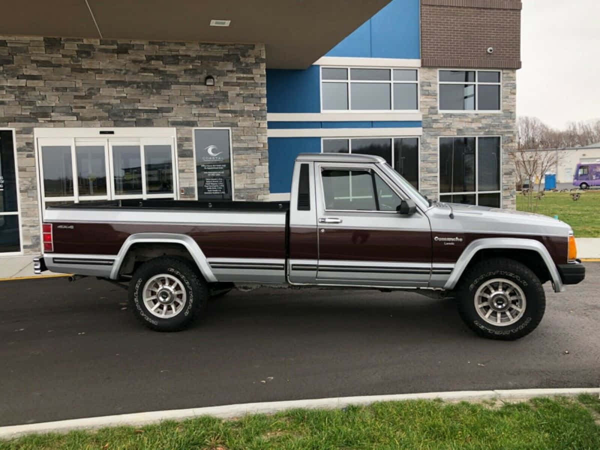 Jeep Comanche 桌布 壁紙 — 一輛栗色和銀色的升高Jeep Comanche皮卡車停在一個現代化的石頭和藍色建築前，天氣陰沉。