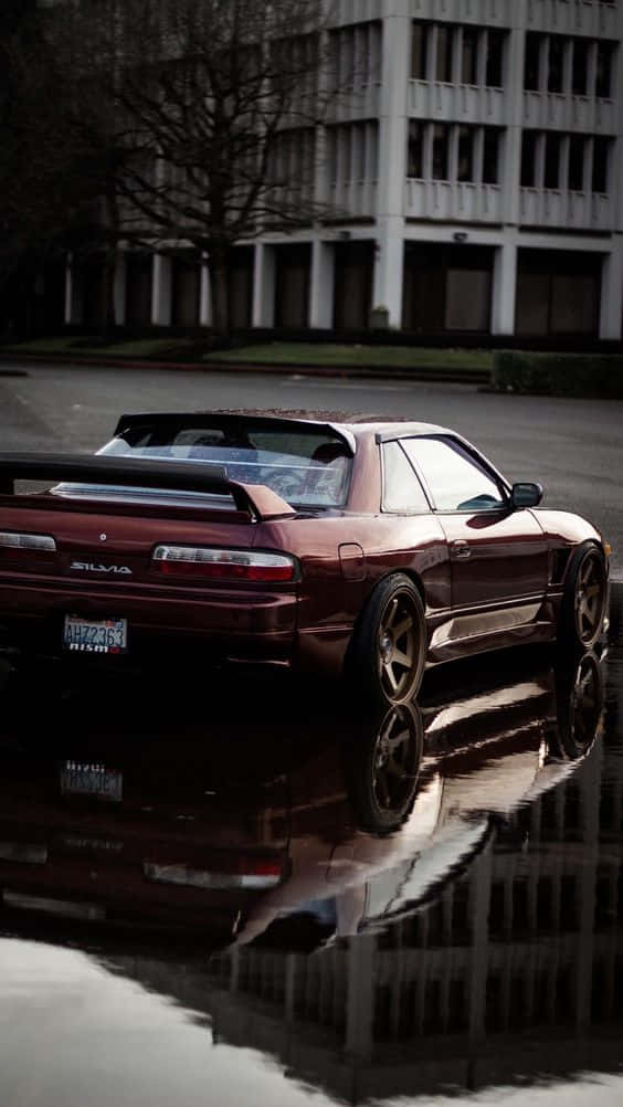 Nissan Silvia S13 桌布 壁紙 — 一輛栗色日產Silvia跑車在濕漉漉的城市街道上的雨水坑中倒映，背景是一座現代建築。