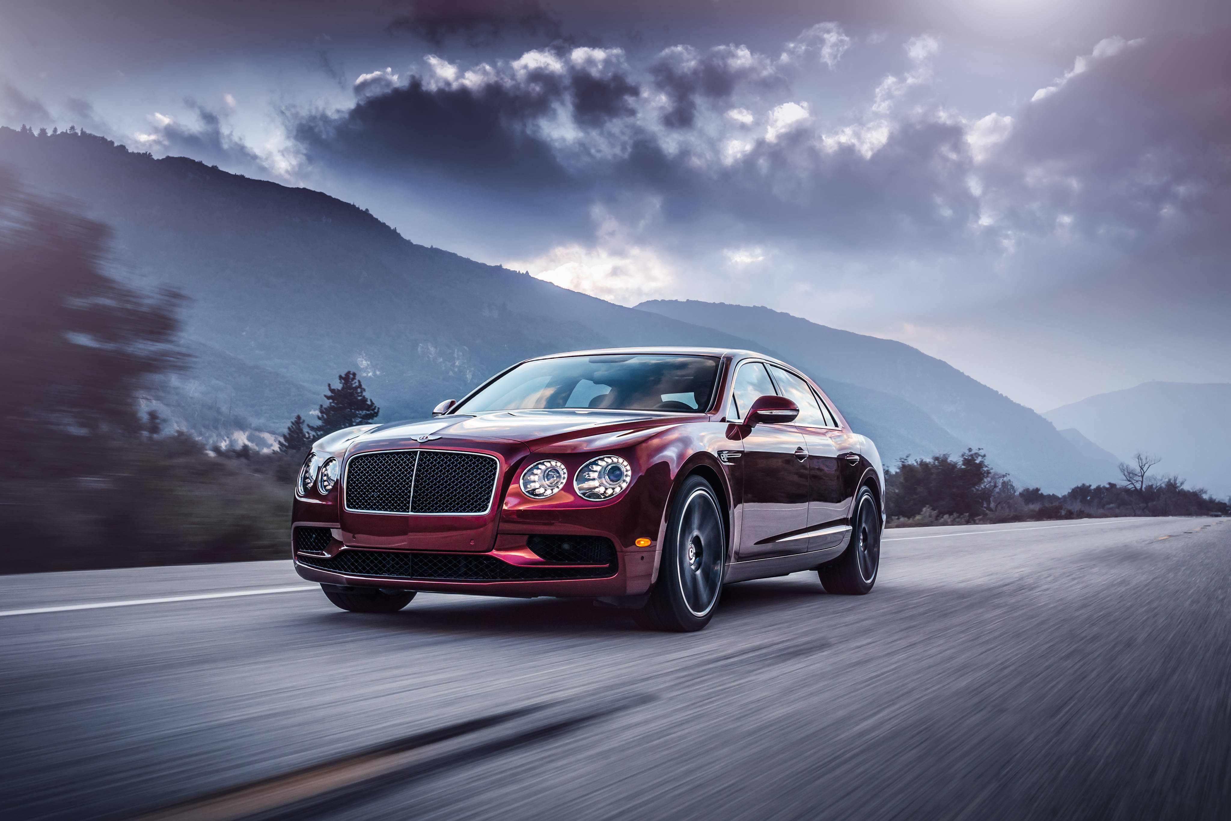賓利 桌布 壁紙 — 一輛栗色賓利 Flying Spur 在戲劇性的多雲天空下沿山路疾馳。