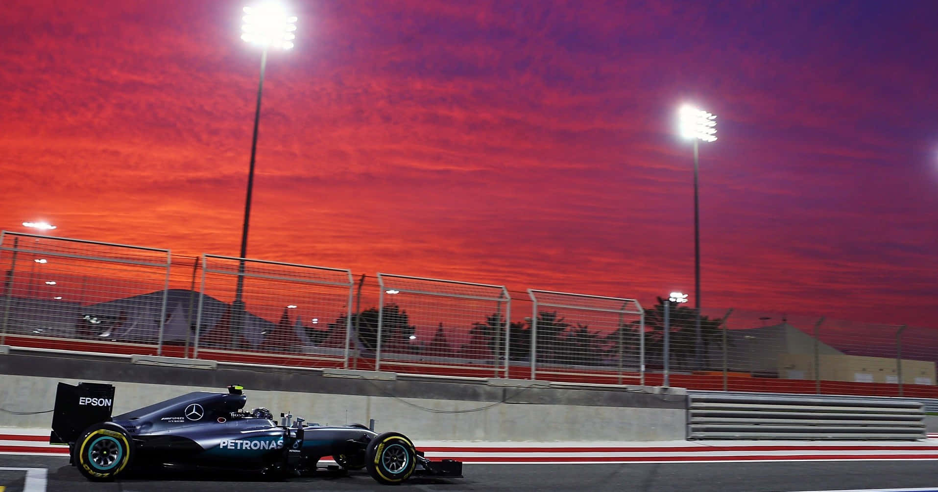 F1 2016 桌布 壁紙 — 一輛梅賽德斯一級方程式賽車在戲劇性的紅色和紫色夕陽天空下，在體育場燈光照亮的賽道上奔馳。