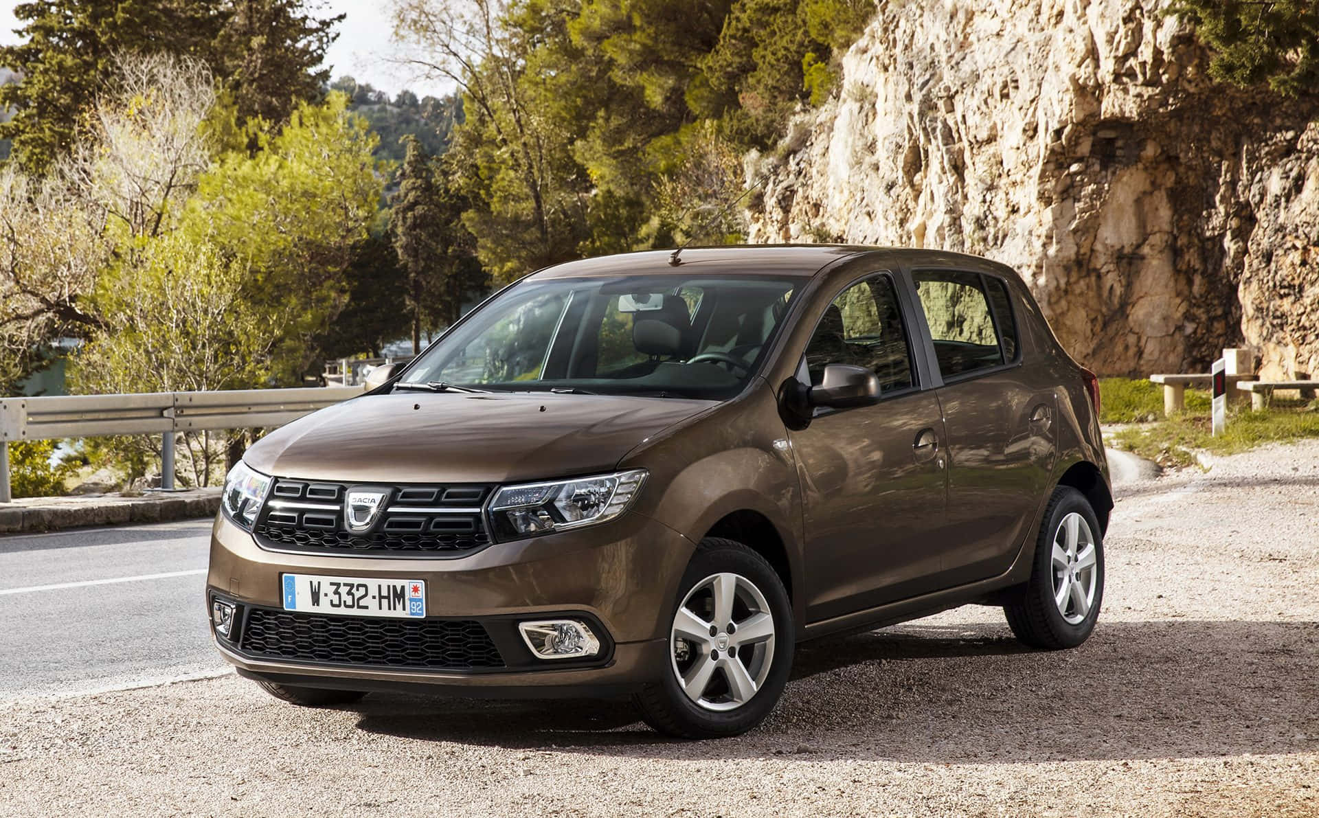 Dacia 桌布 壁紙 — 一輛棕色Dacia Sandero停放在碎石路邊，旁邊是岩石懸崖和綠樹，在明亮的日光下。