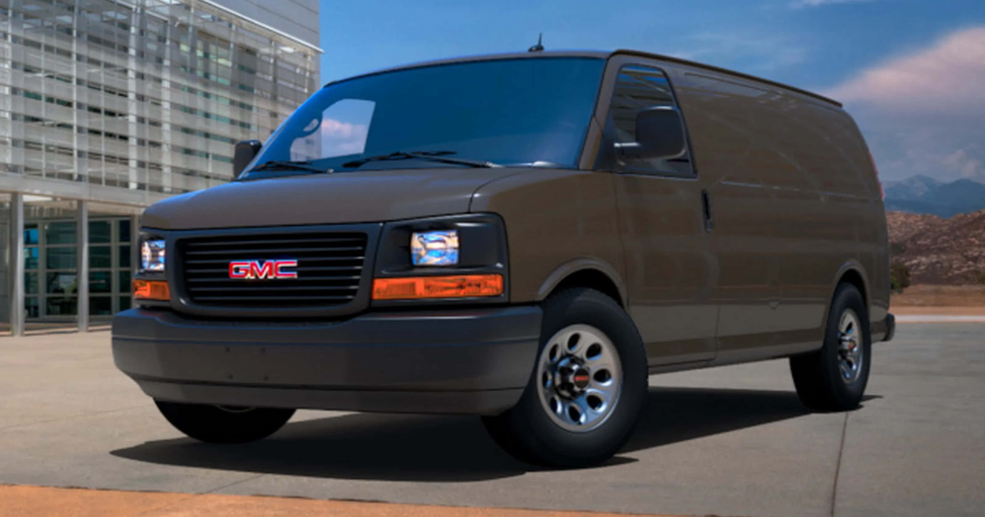 Gmc Savana 桌布 壁紙 — 一輛棕色GMC貨車停在人行道上，背景有現代建築和遠處的山脈，天空清澈湛藍。