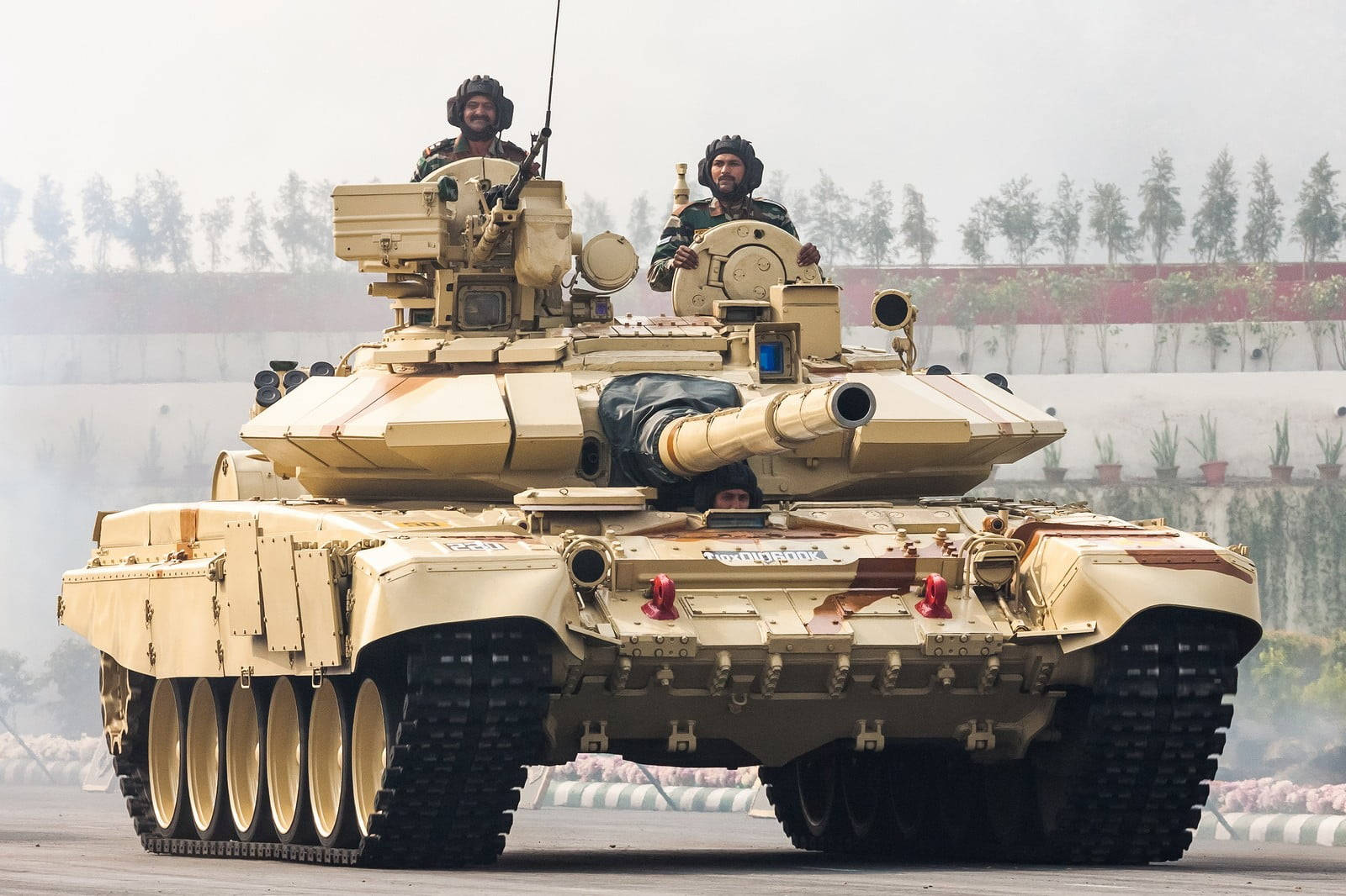印度軍隊 桌布 壁紙 — 一輛棕褐色裝甲坦克，頂部有士兵，在鋪砌的軌道上向前移動，身後留下煙霧。