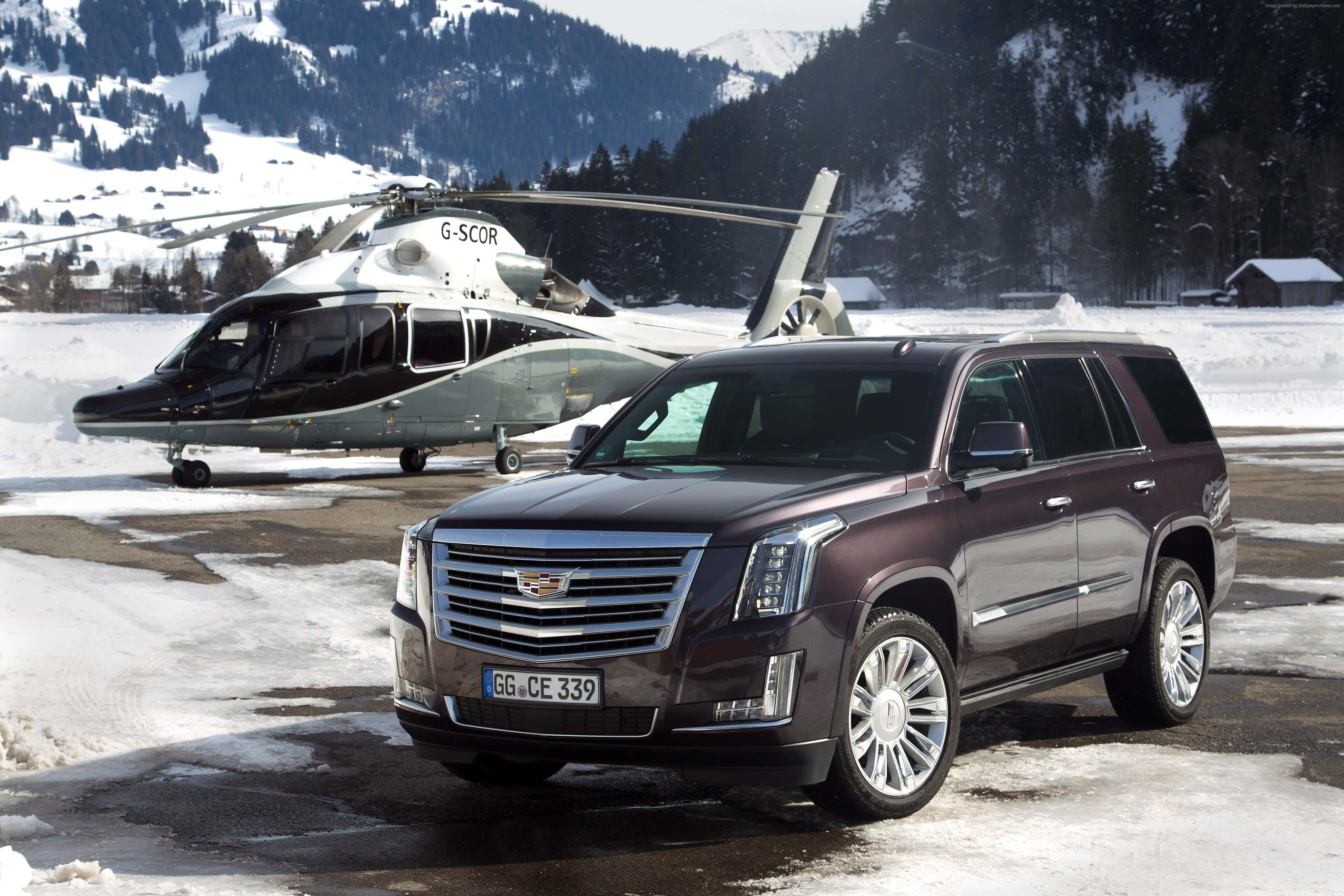 Cadillac Escalade 桌布 壁紙 — 一輛棗紅色 Cadillac Escalade SUV 停放在積雪的停機坪上，旁邊有一架黑白相間的直升機，背景是白雪覆蓋的山脈。
