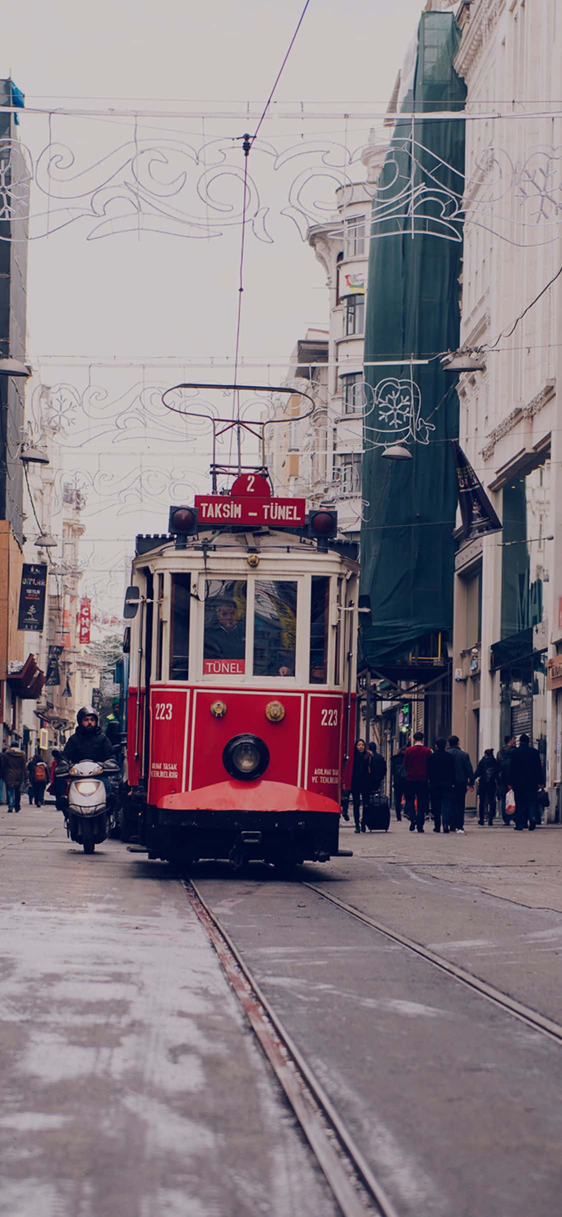 城市 桌布 壁紙 — 一輛標有Taksim-Tunel的復古紅色電車在伊斯坦堡濕漉漉的鵝卵石街道上行駛，上方裝飾著白色串燈。
