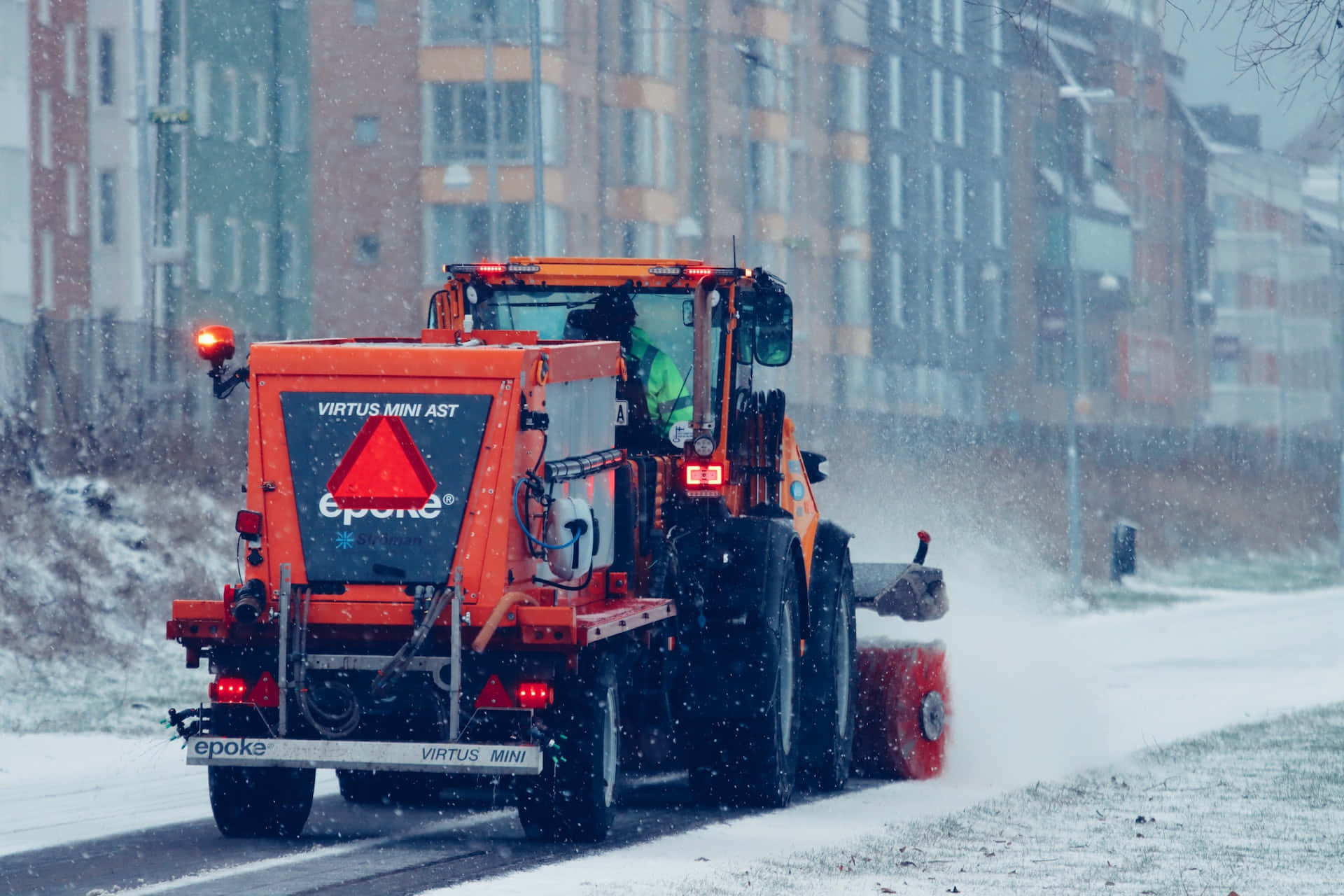 Snowplow 桌布 壁紙 — 一輛橙色鏟雪車在積雪的城市街道上鏟雪，雪花在模糊的公寓大樓背景下飄落。