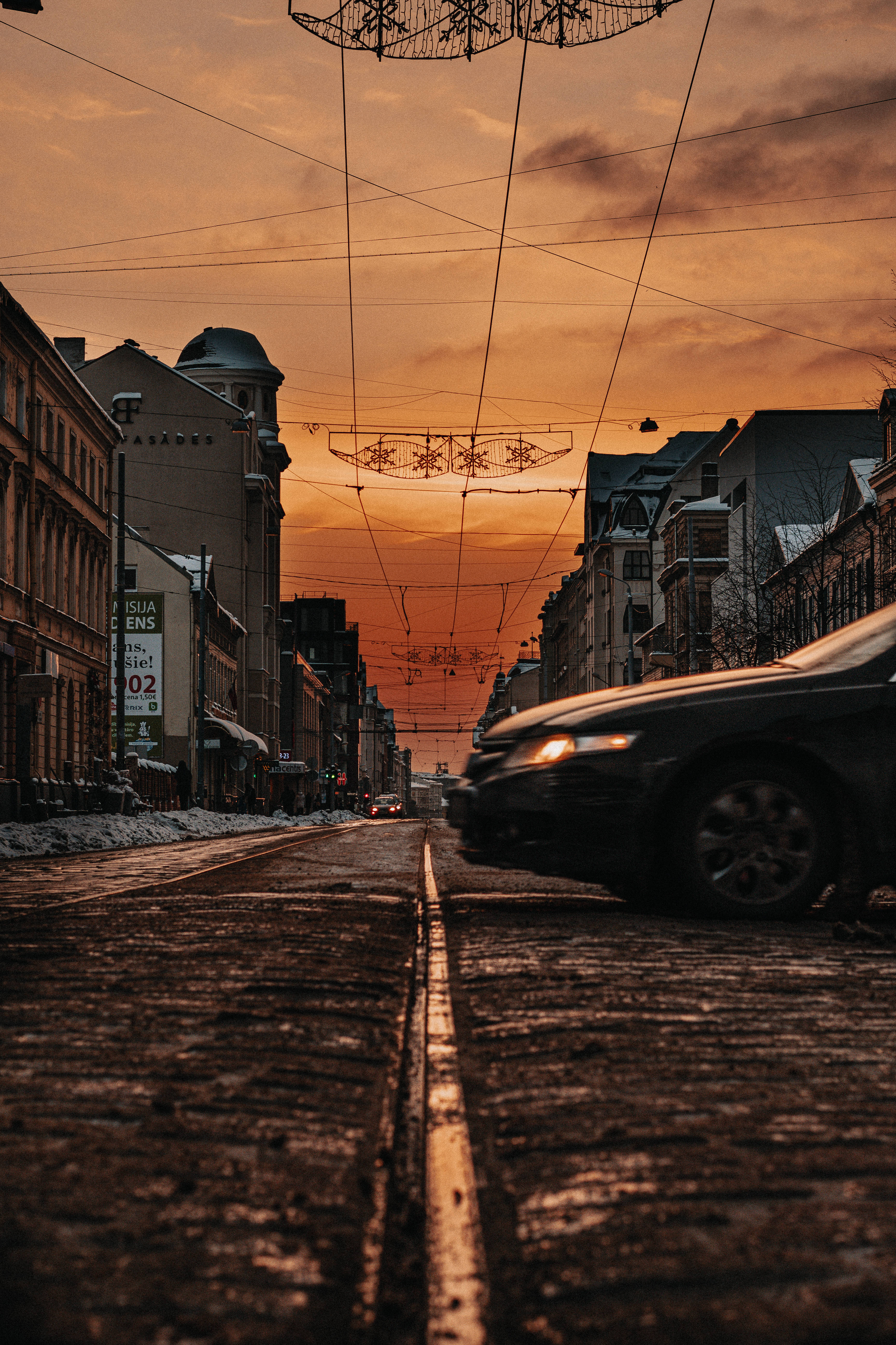 Latvia 桌布 壁紙 — 一輛汽車在日落時分沿著鵝卵石街道疾馳，節日燈飾懸掛在積雪的建築物上方。