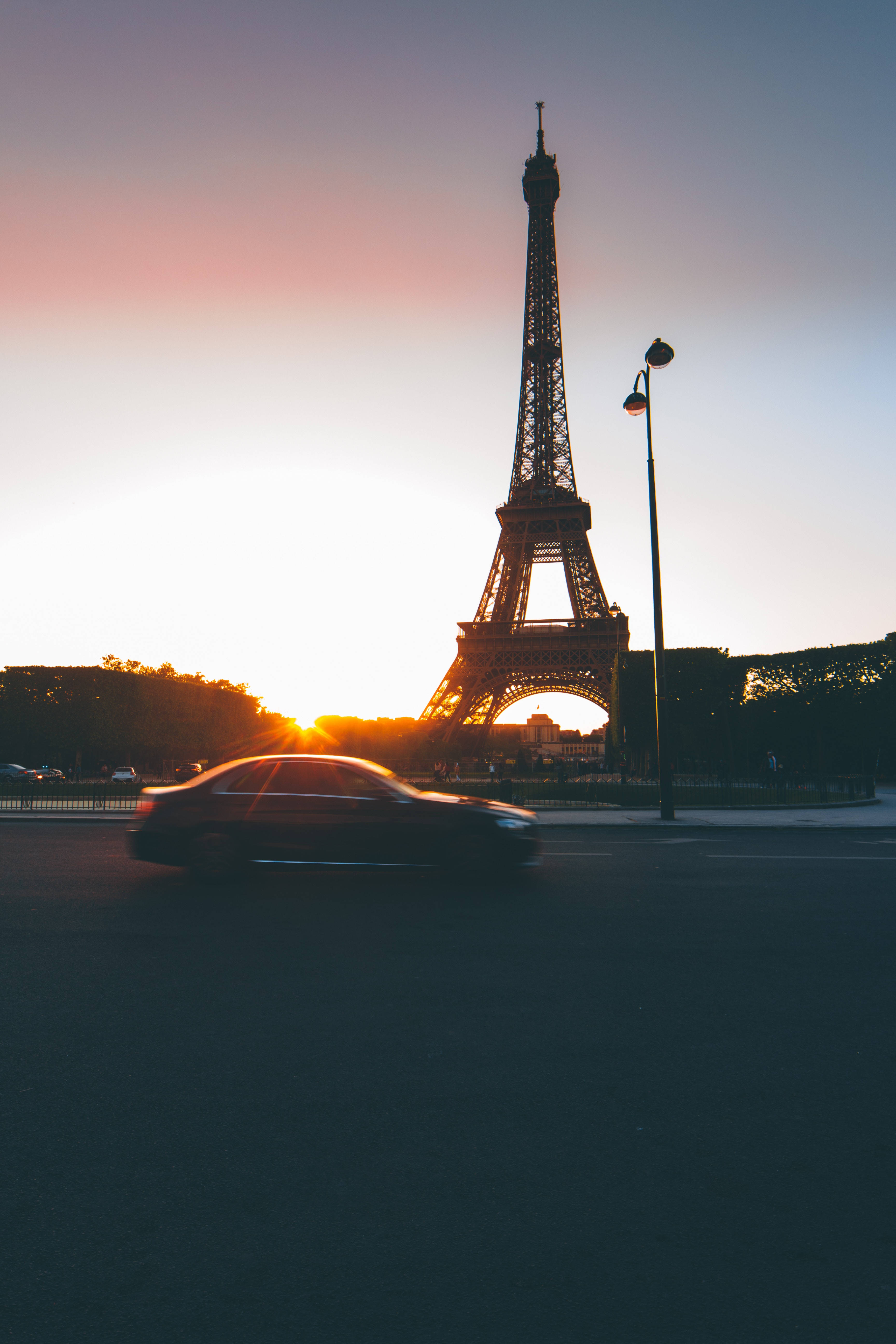 Eiffel Tower 桌布 壁紙 — 一輛汽車在日落時模糊地駛過艾菲爾鐵塔，在柔和的粉紅色和藍色天空中投射出溫暖的金色光芒。