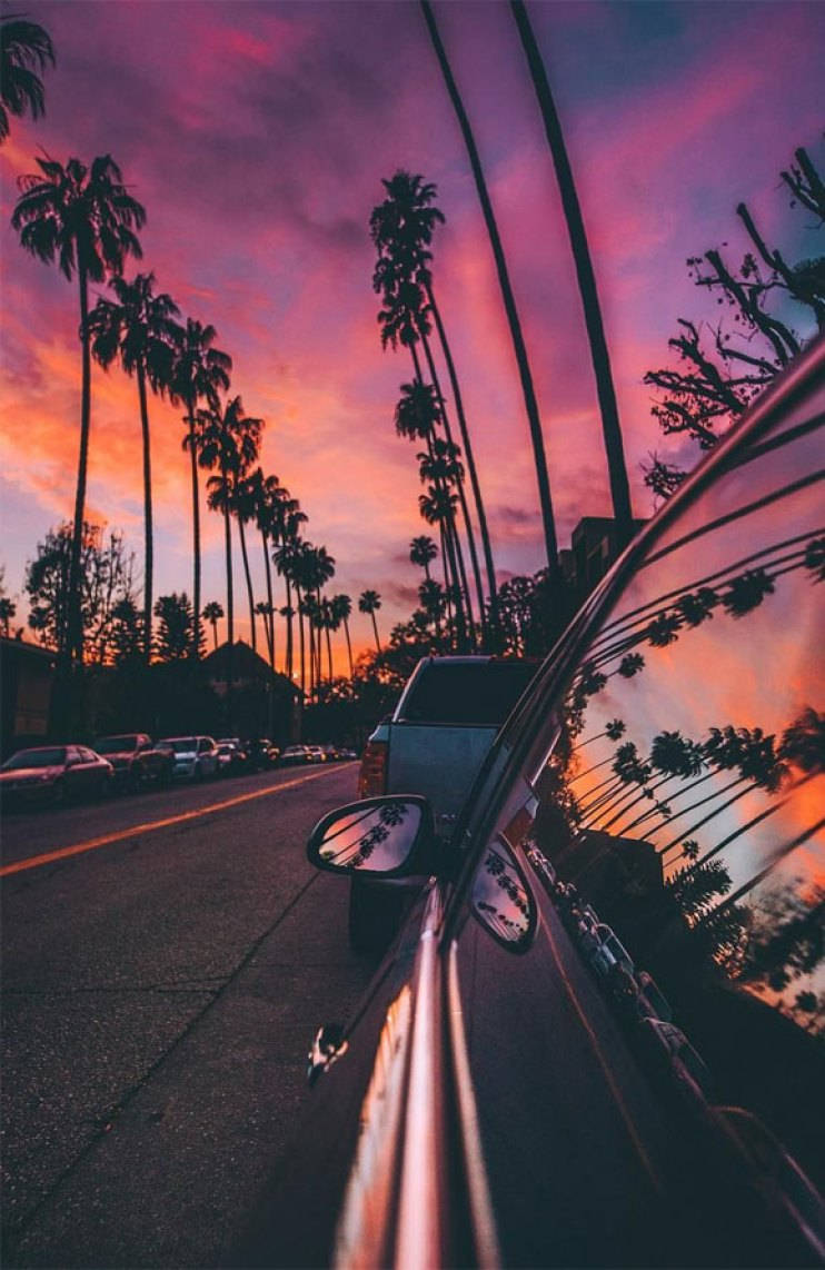 Pink Sunset iPhone 桌布 壁紙 — 一輛汽車沿著棕櫚樹成蔭的街道行駛，熾熱的夕陽將天空染成鮮豔的粉紅色和橙色。