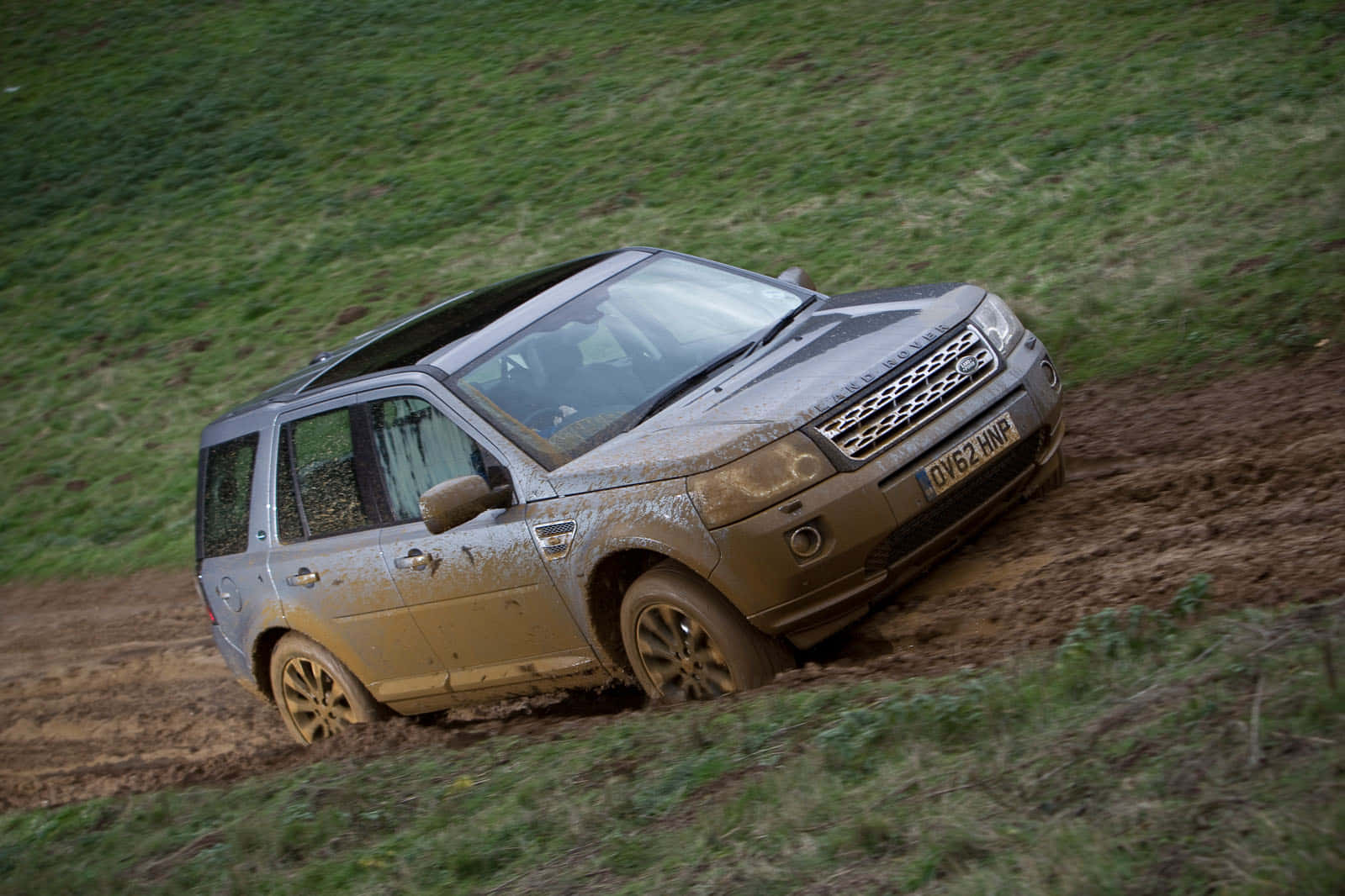 Land Rover Freelander 桌布 壁紙 — 一輛沾滿泥濘的銀色SUV駛上長滿草的山坡，在濕潤的泥土上留下深深的輪胎痕跡。