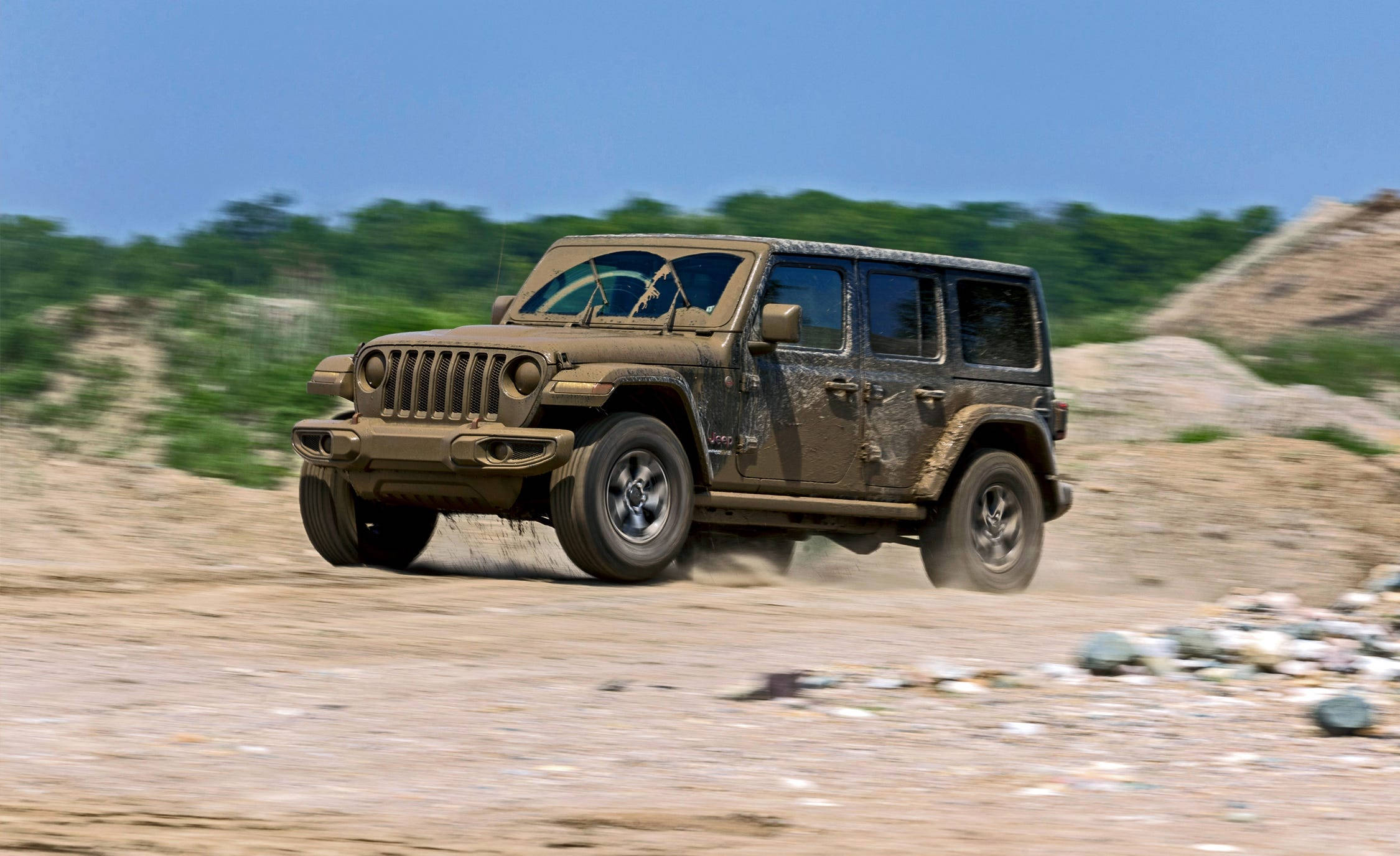 Wrangler 桌布 壁紙 — 一輛泥濘的Jeep Wrangler在岩石沙漠地形上高速行駛，在晴朗的藍天下揚起灰塵。